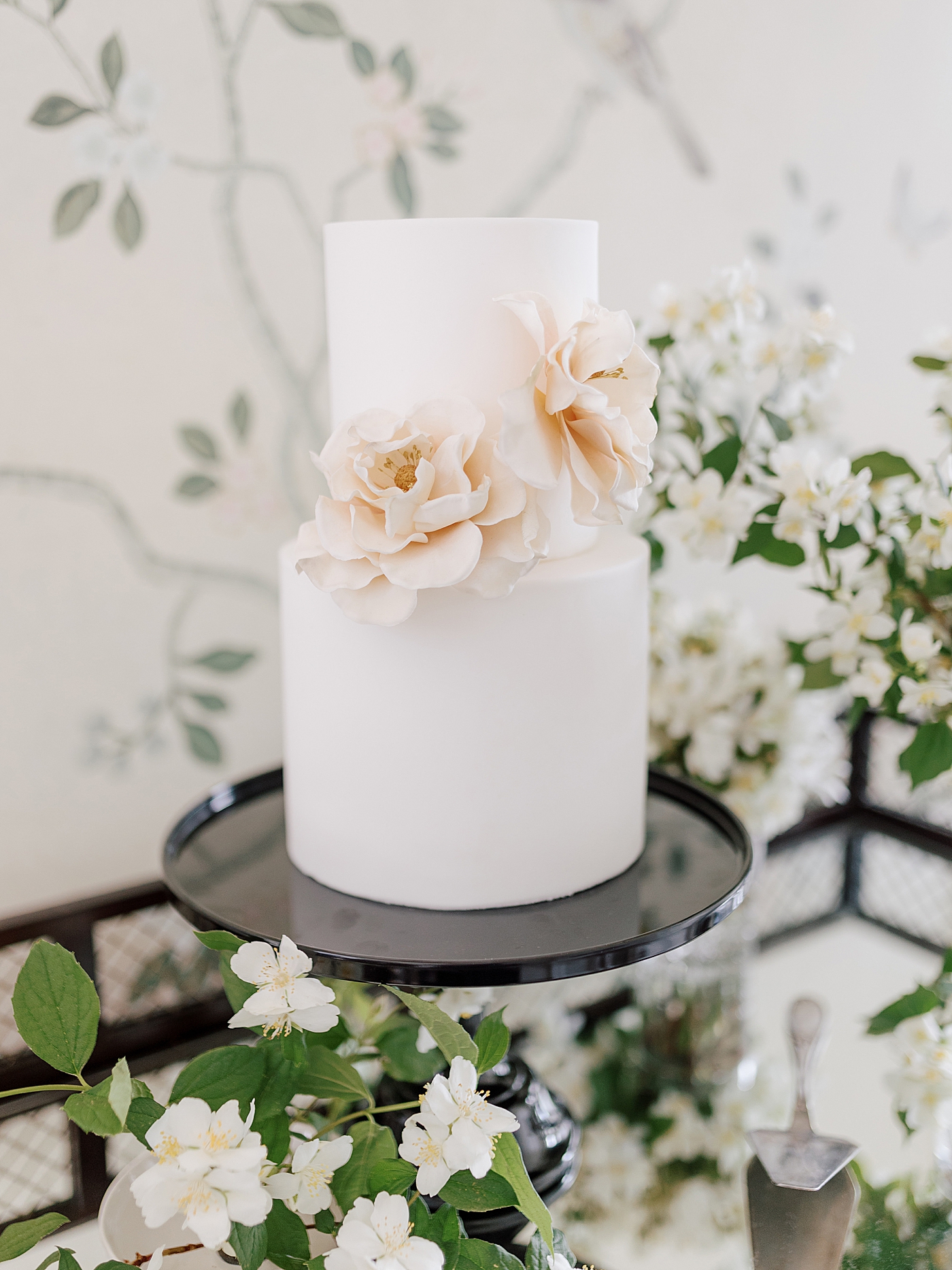 White wedding cake with pink sugar flowers in front of floral wallpaper | Photo by Hope Helmuth Photography