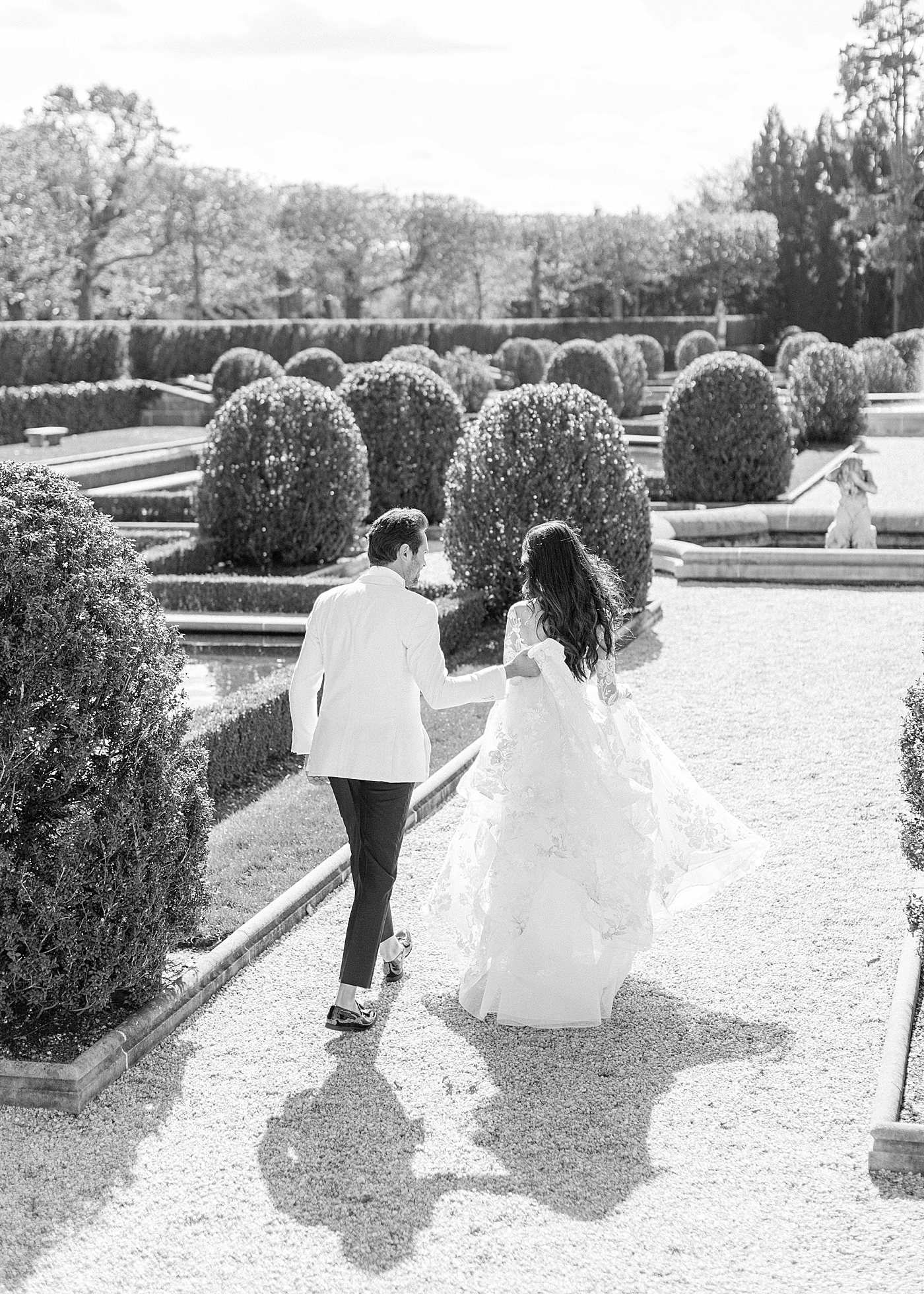 Black and white image of a bride and groom walking away from the camera in a European garden during Oheka castle wedding | Image by Hope Helmuth Photography