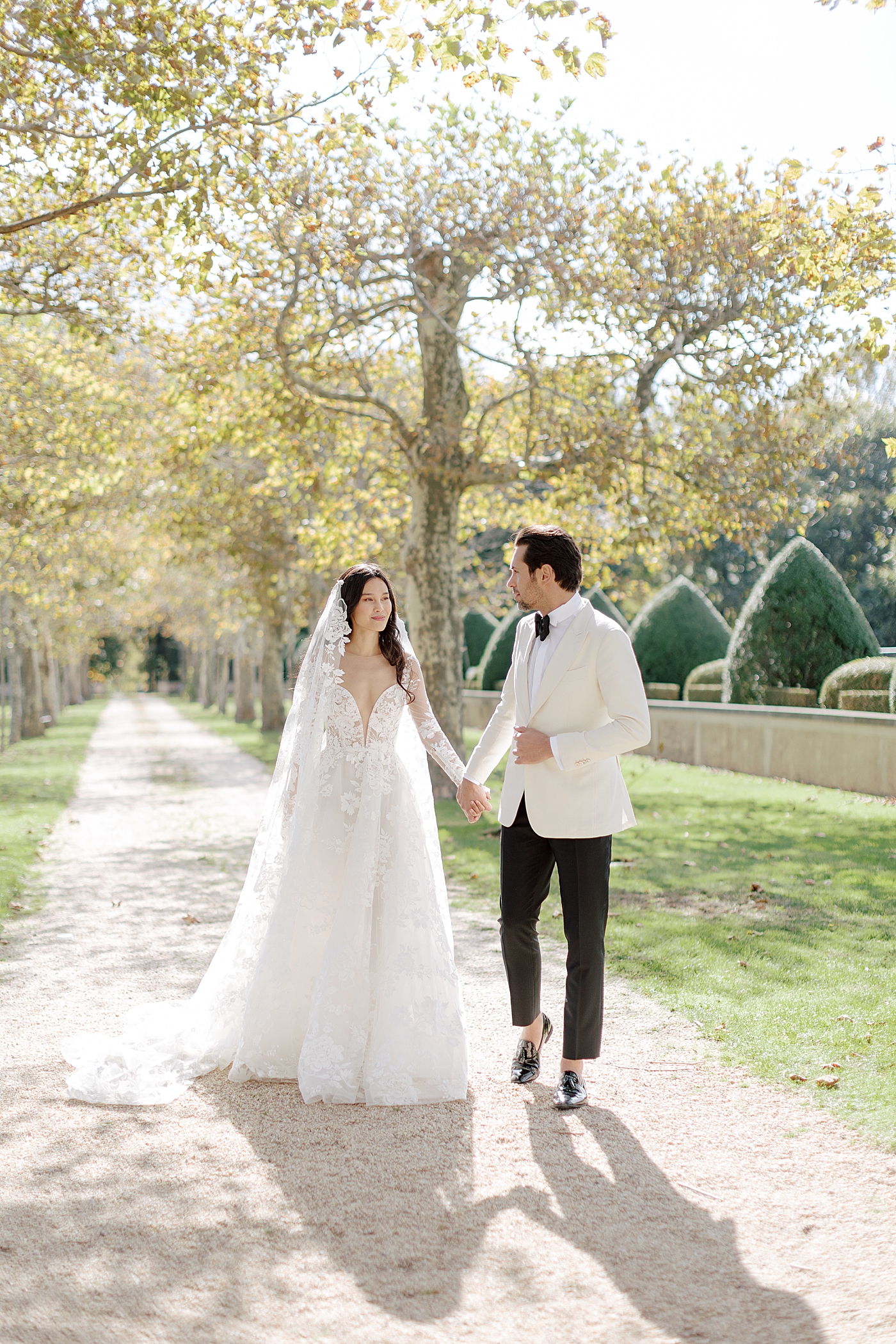 Portrait of bride and groom walking at the end of a tree-lined driveway during Oheka castle wedding | Image by Hope Helmuth Photography