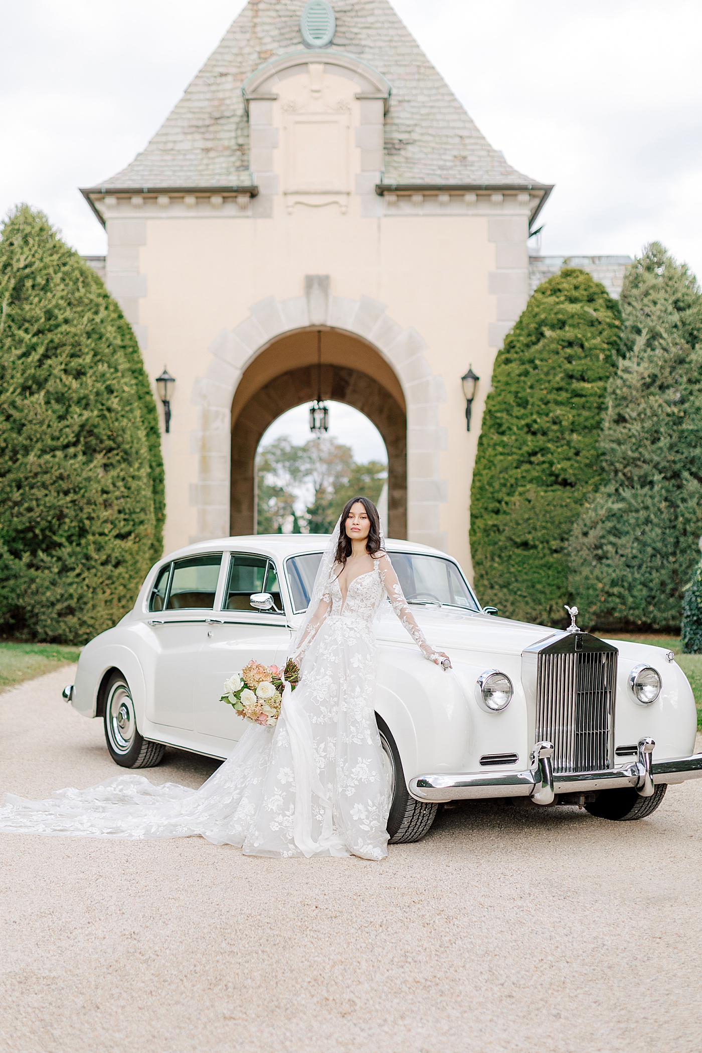 Bride leaning on a classic car in a gravel driveway during Oheka castle wedding | Image by Hope Helmuth Photography