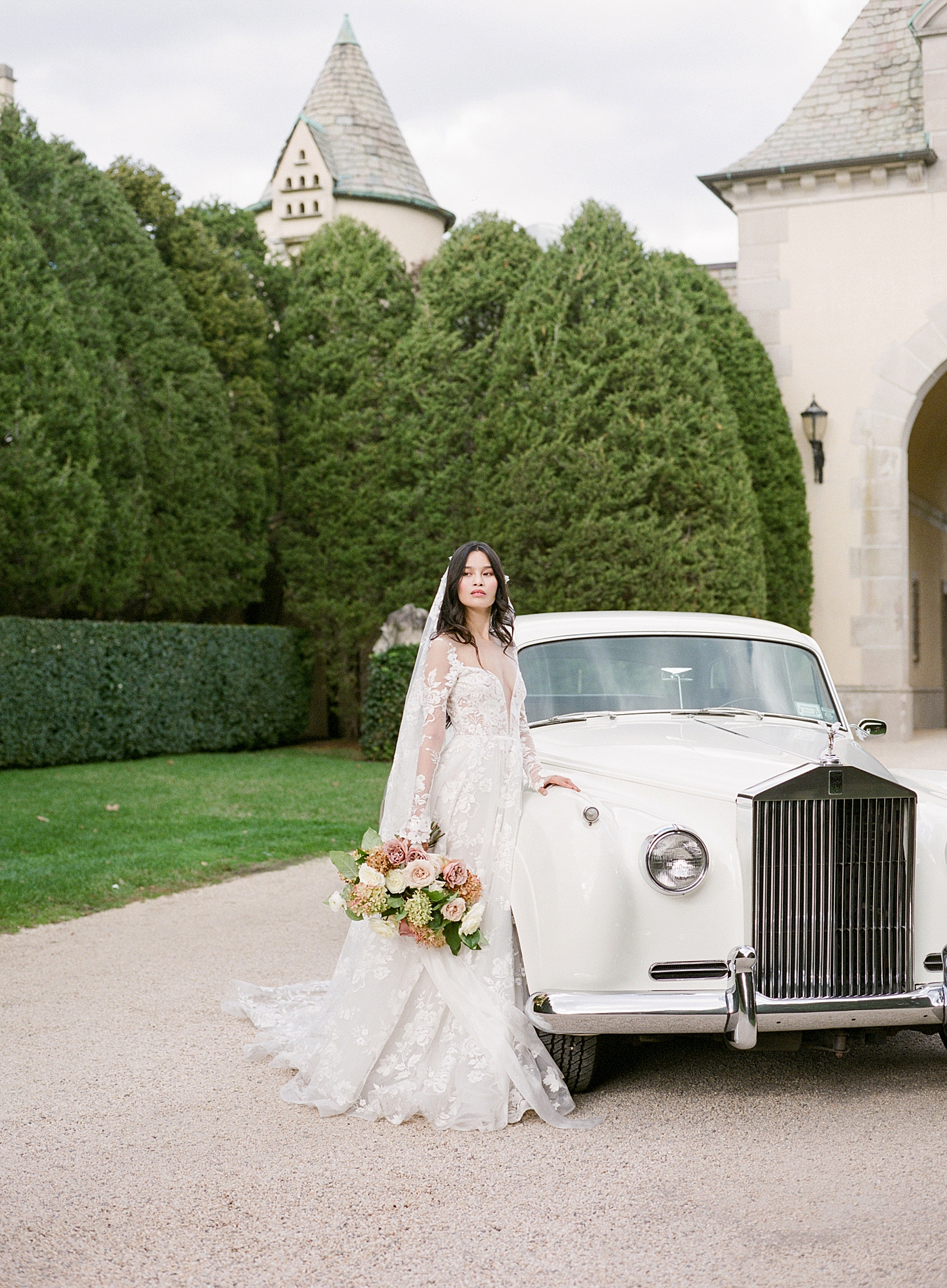 Bride leaning on a classic car in a gravel driveway during Oheka castle wedding | Image by Hope Helmuth Photography