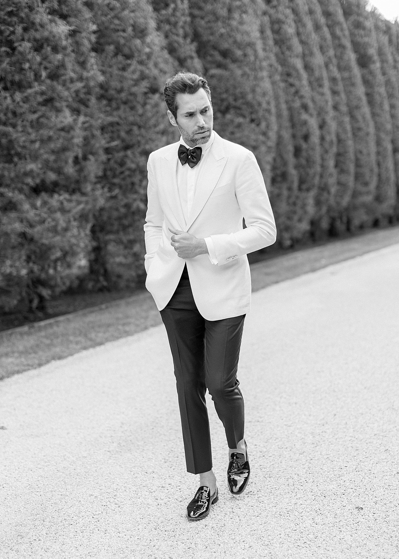 Black and white photo of a groom in a white and black tux walking down a gravel driveway | Image by Hope Helmuth Photography
