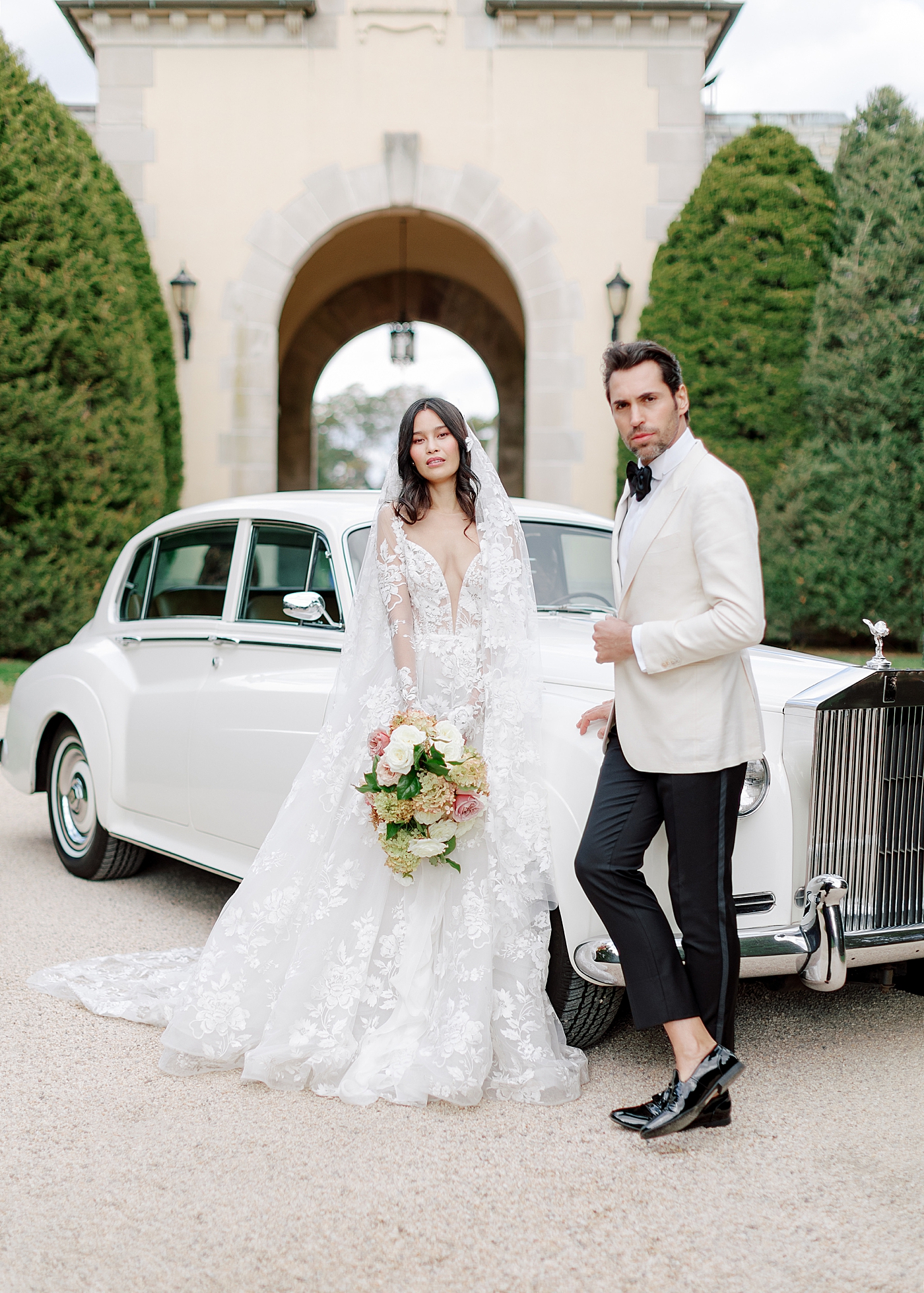 Bride and groom leaning on a classic car in a gravel driveway during Oheka castle wedding | Image by Hope Helmuth Photography