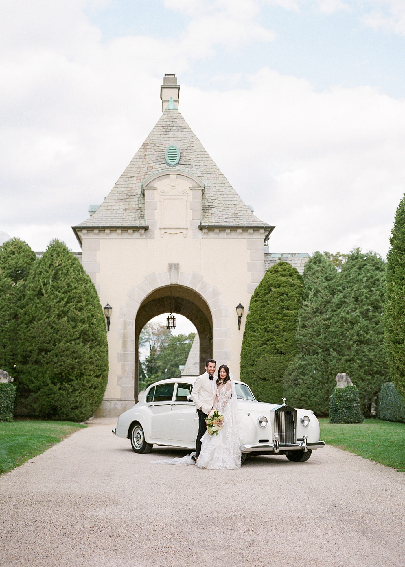 Bride and groom leaning on a classic car in a gravel driveway during Oheka castle wedding | Image by Hope Helmuth Photography