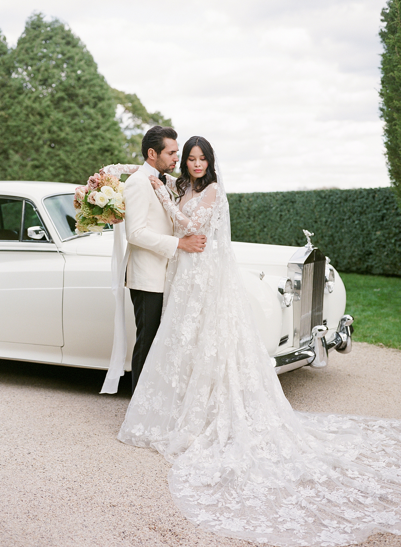 Bride and groom embracing by a classic car in a gravel driveway during Oheka castle wedding | Image by Hope Helmuth Photography