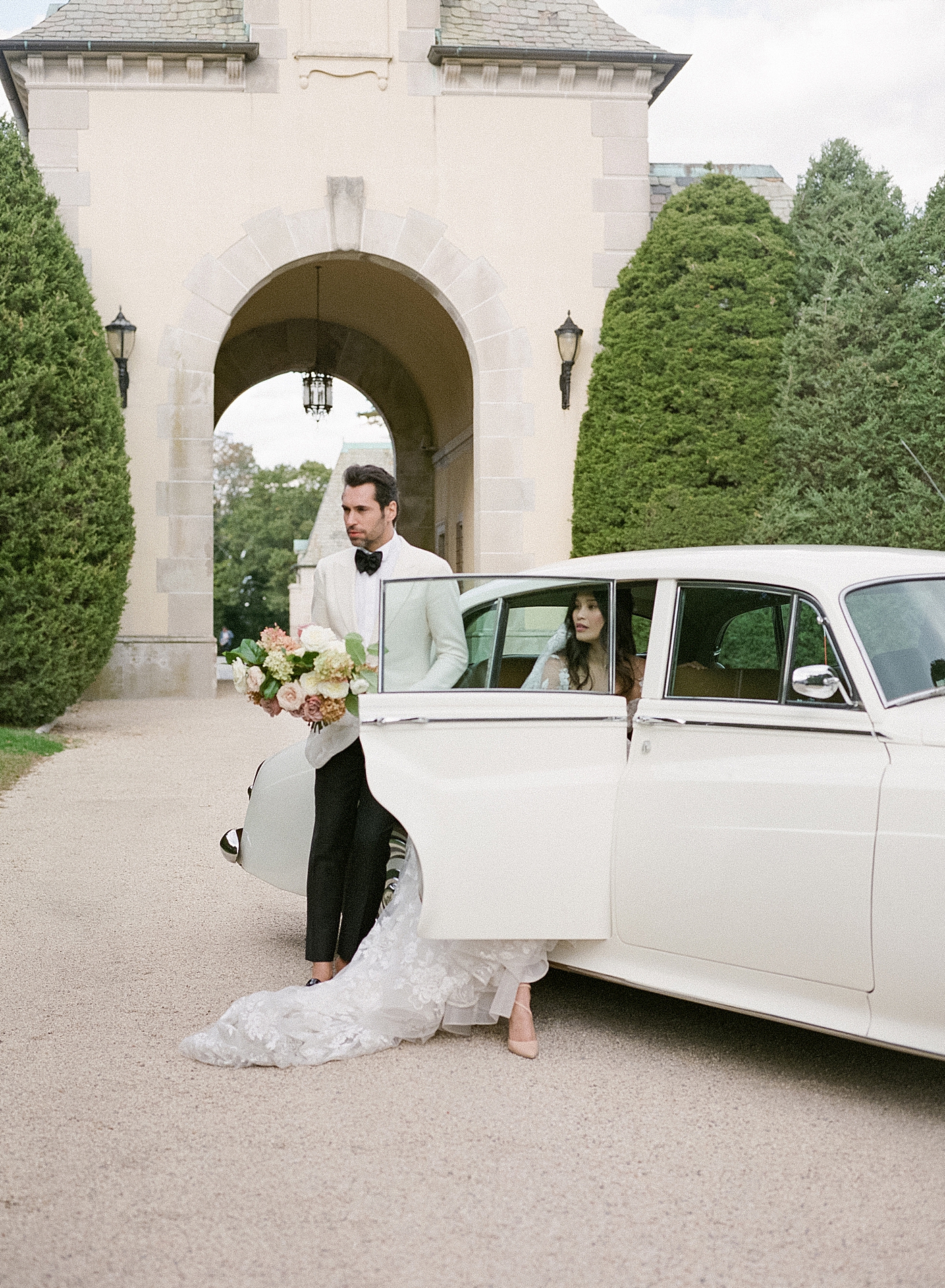 Bride and groom getting into a classic car in a gravel driveway during Oheka castle wedding | Image by Hope Helmuth Photography