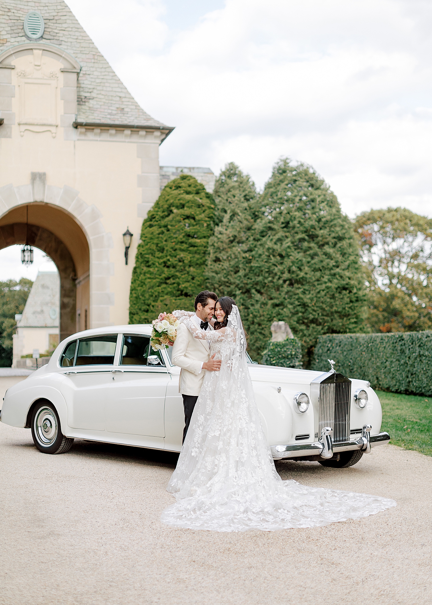 Bride and groom embracing by a classic car in a gravel driveway during Oheka castle wedding | Image by Hope Helmuth Photography