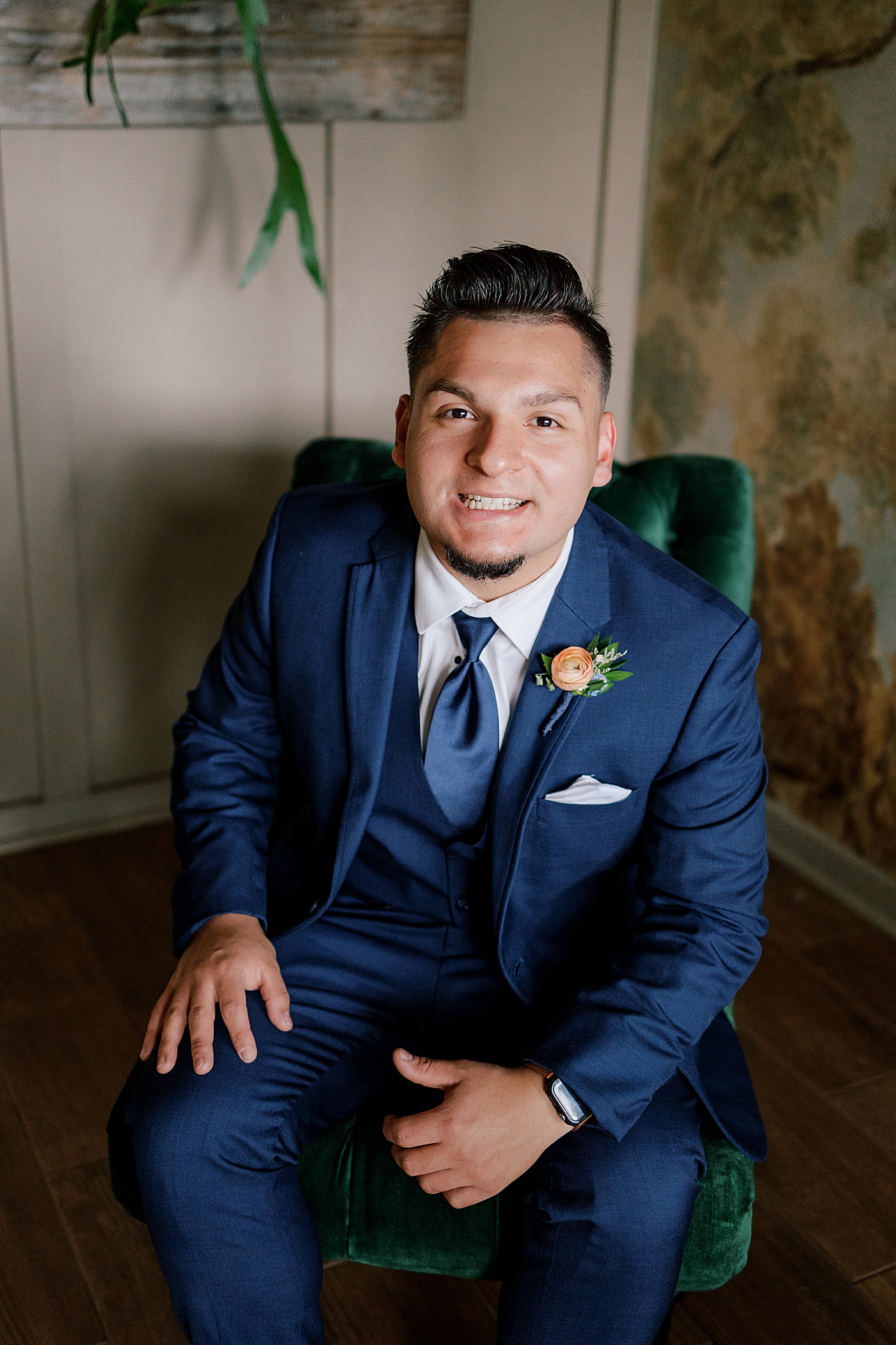 Groom in a navy suit sitting in a green chair smiling | Image by Hope Helmuth Photography 