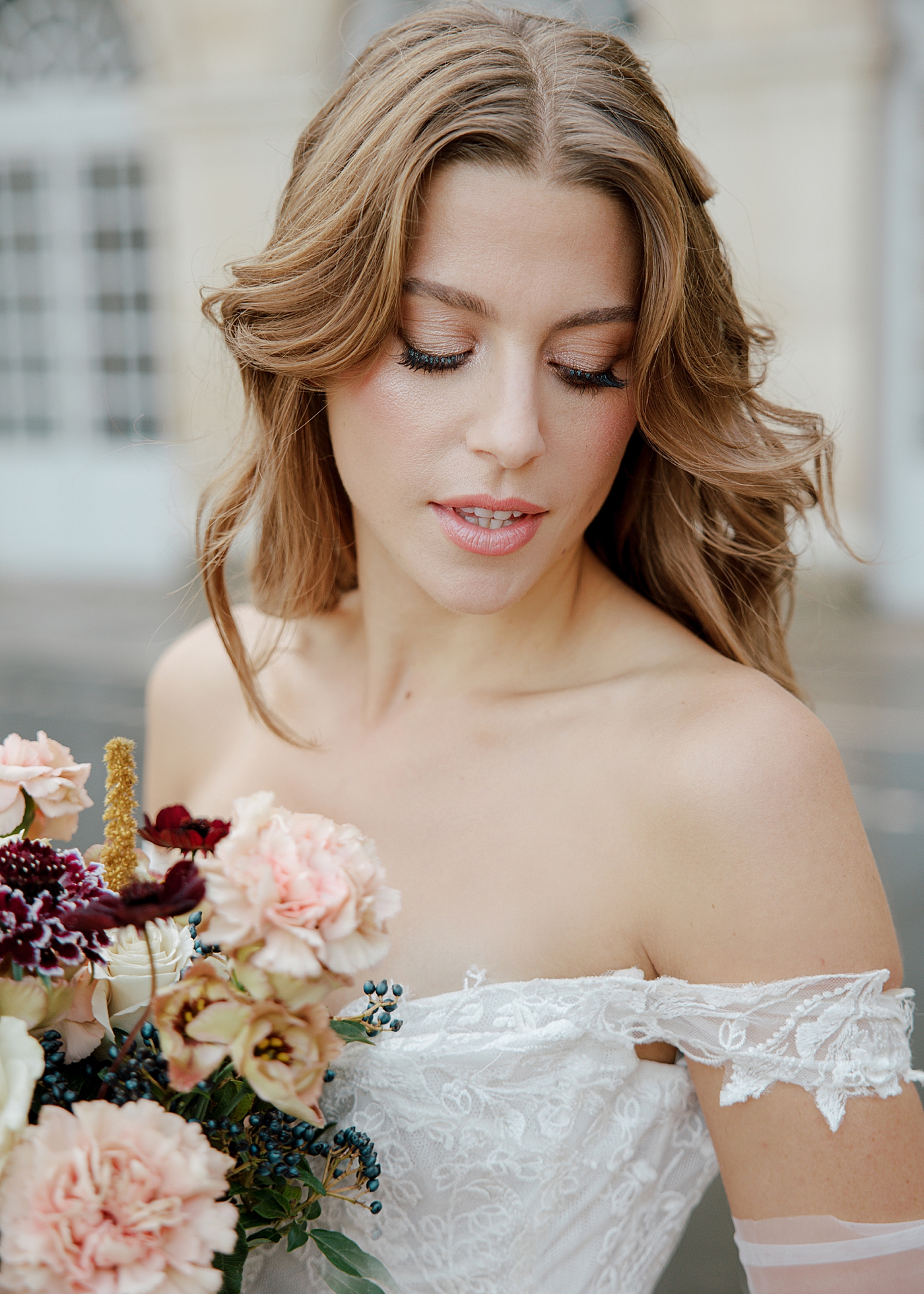 Close up of bride in an off the shoulder dress and white sheer gloves holding a wedding bouquet while looking down over her shoulder | Image by Hope Helmuth Photography
