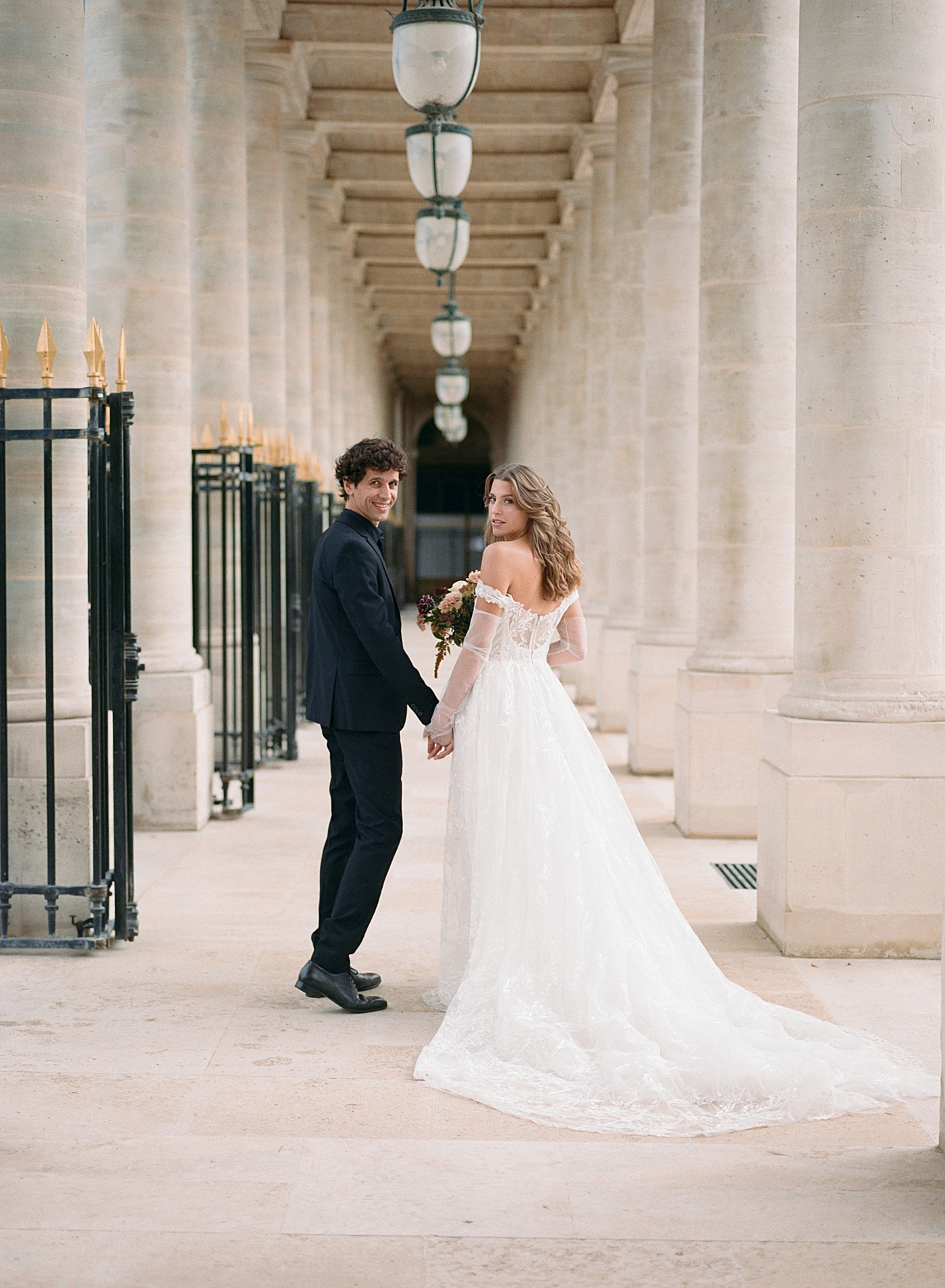 Image of a bride and groom holding hands and smiling back at the camera while walking away down an outdoor, sunlit, concrete corridor | Image by Hope Helmuth Photography