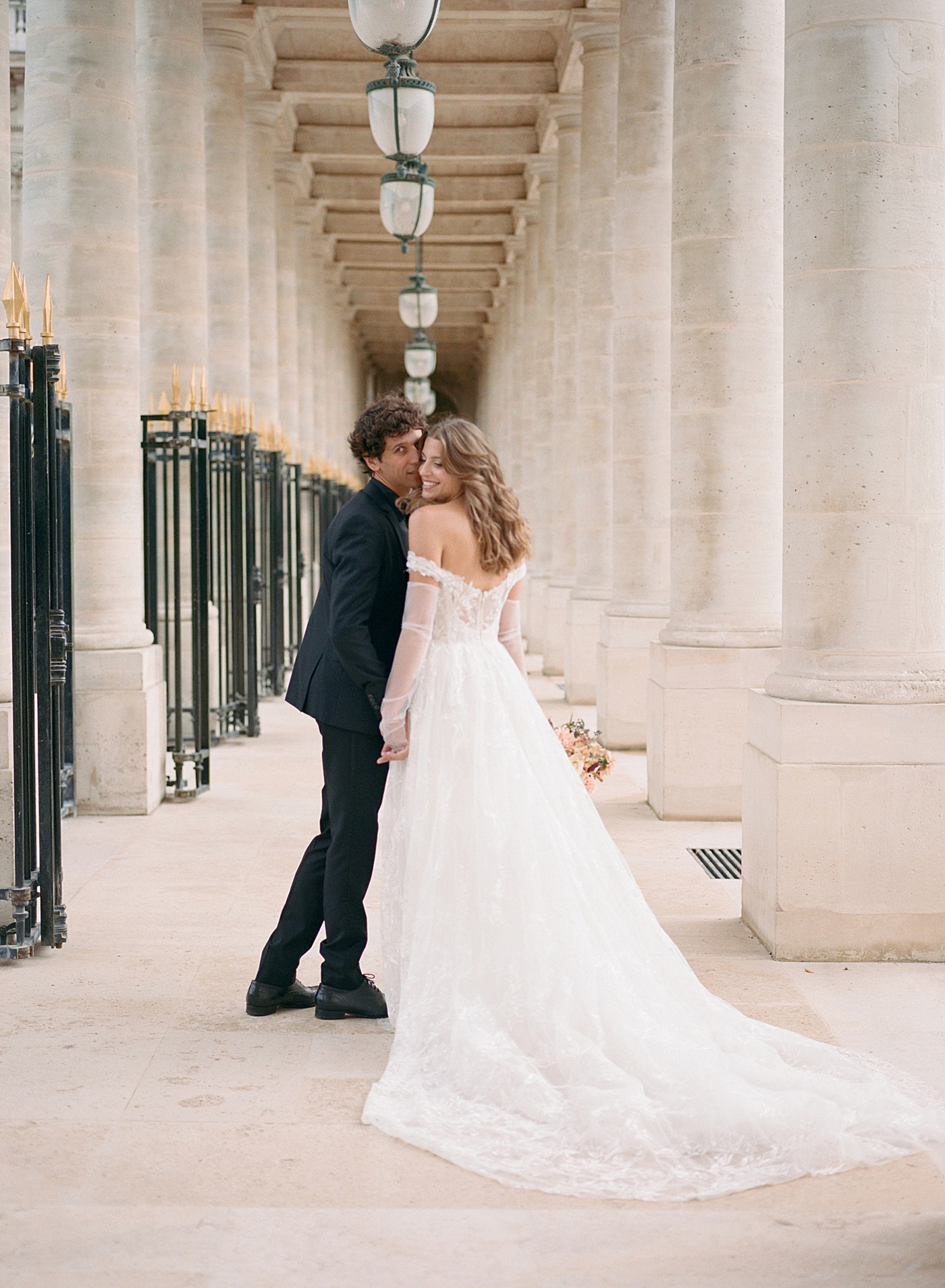 Image of a bride and groom holding hands, kissing, and smiling back at the camera while walking away down an outdoor, sunlit, concrete corridor | Image by Hope Helmuth Photography