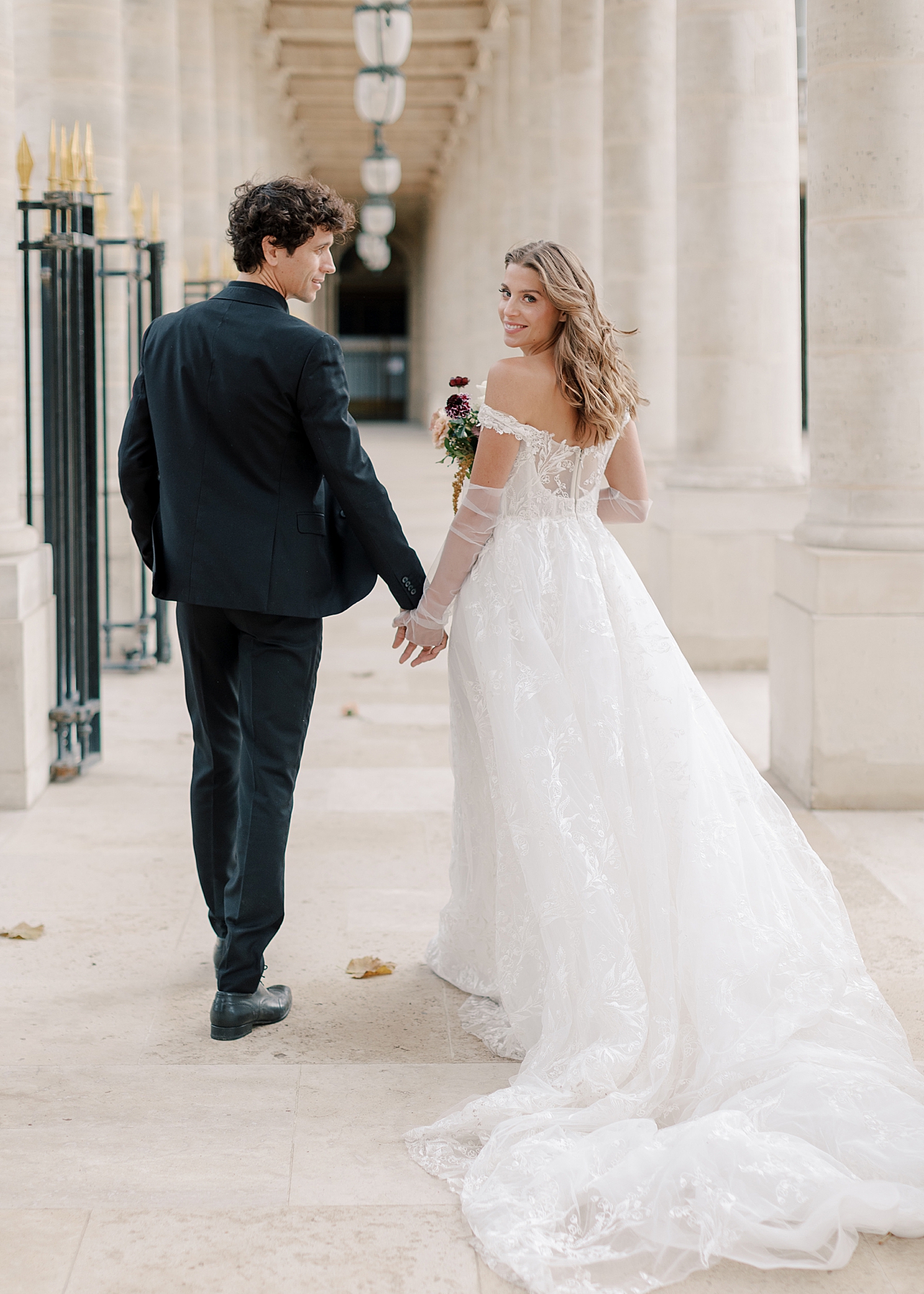 Image of a bride and groom holding hands and walking away in an outdoor, sunlit, concrete corridor| Image by Hope Helmuth Photography