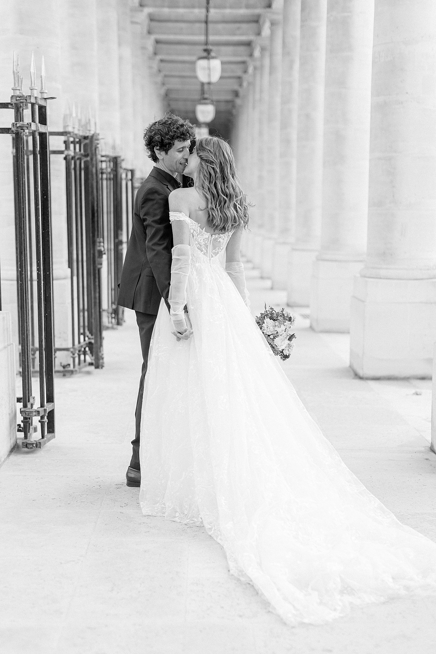 Black and white image of a bride and groom embracing and kissing in an outdoor, sunlit, concrete corridor | Image by Hope Helmuth Photography