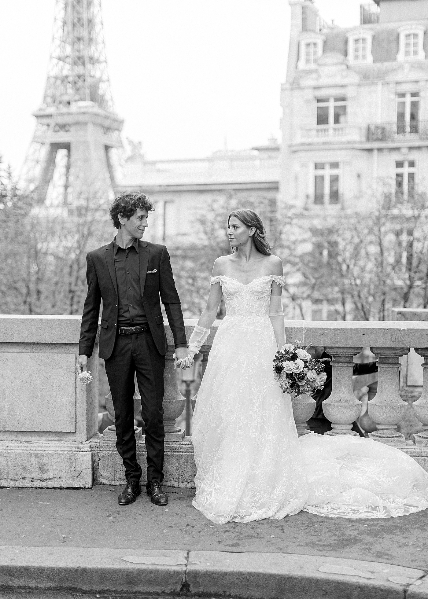 Black and white image of a bride and groom holding hands and walking towards the camera in Paris with the Eiffel tower in the background | Image by Hope Helmuth Photography