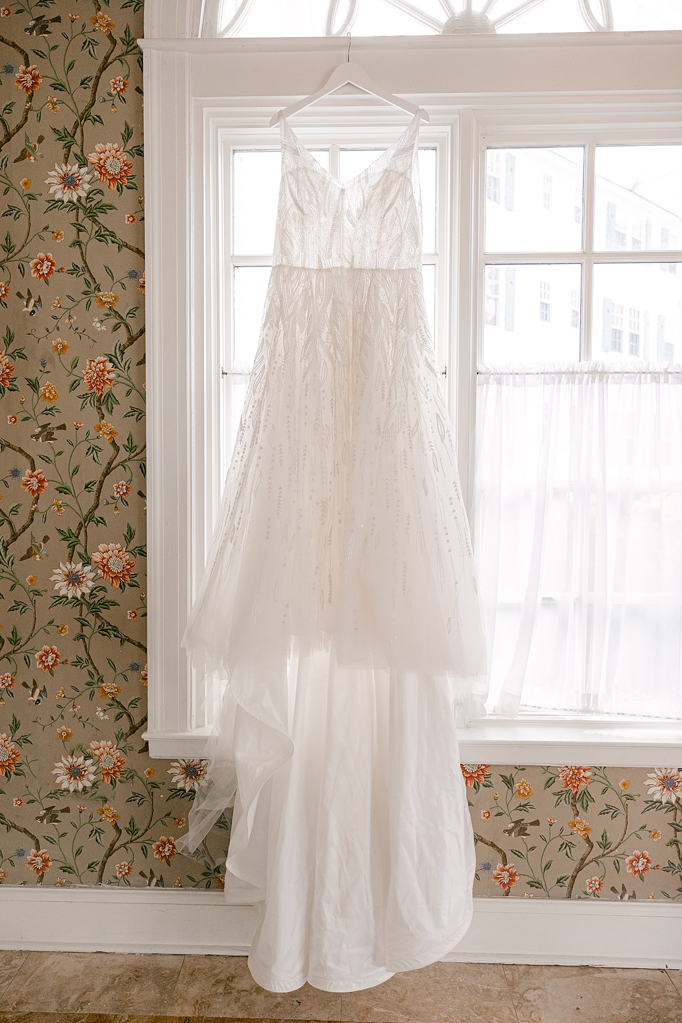 Sheer wedding dress hanging in front of a window with a glimpse of floral wall paper | Image by Hope Helmuth Photography