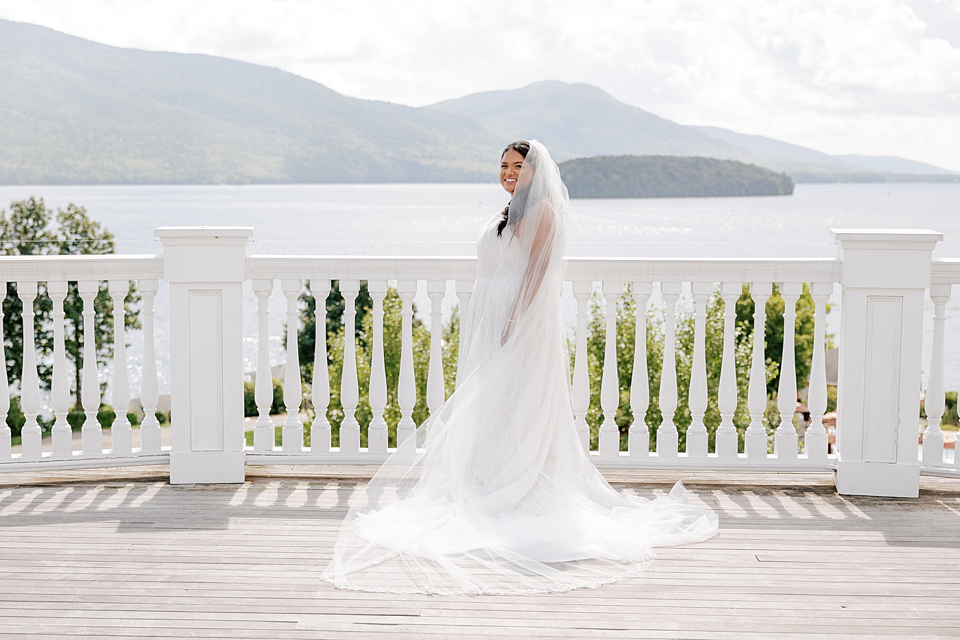 Bridal portrait of a bride on a deck with white railing and water and mountains in the background | Image by Hope Helmuth Photography