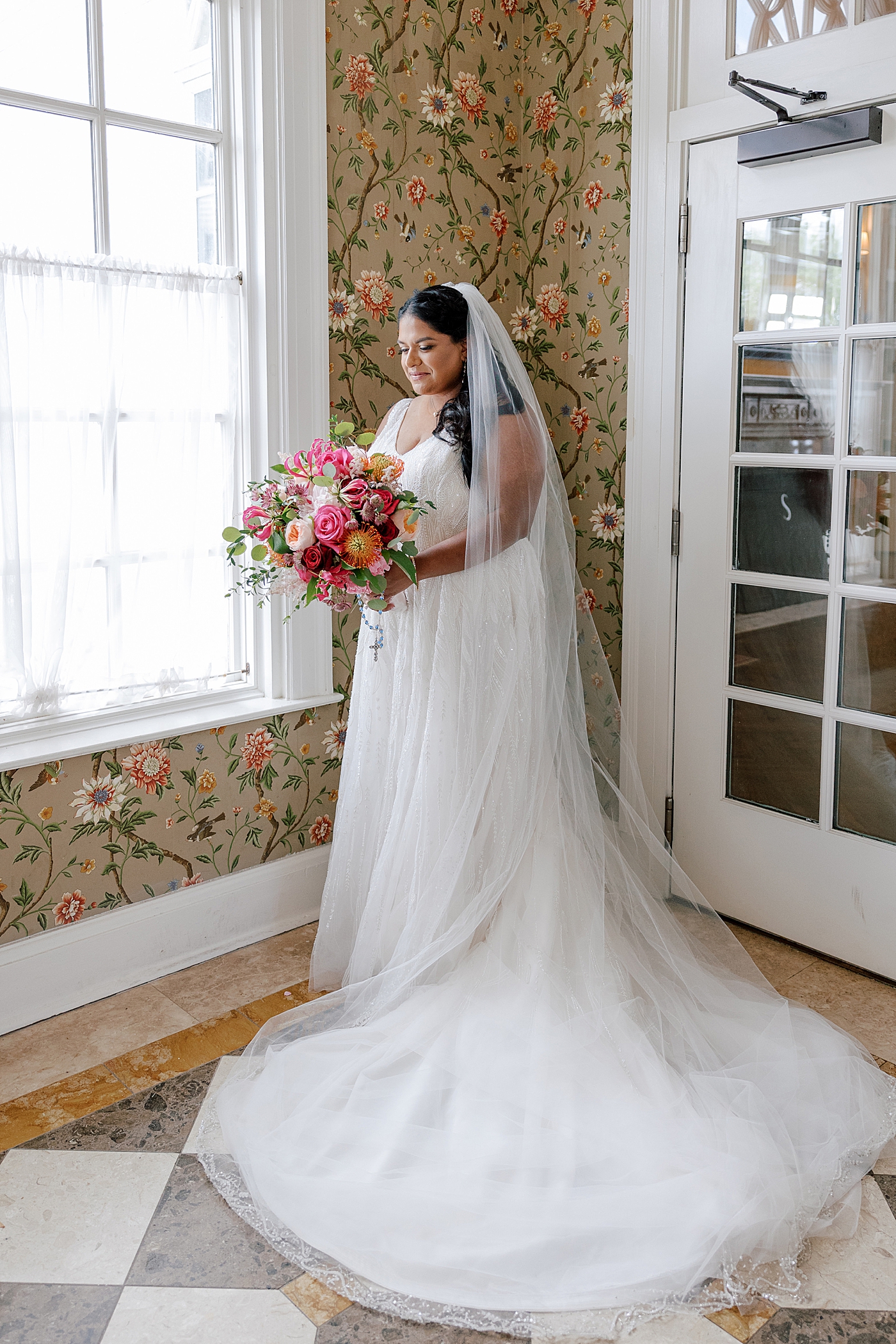 Bride posing indoors with floral wallpaper looking down at her bouquet | Image by Hope Helmuth Photography