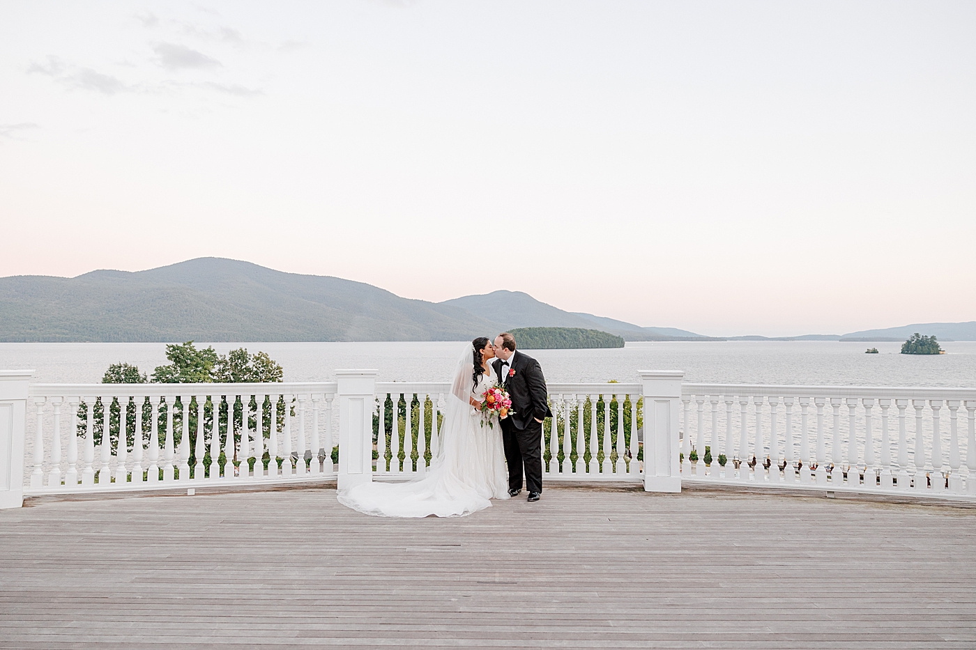 Bride and groom kissing on a deck with water and mountains in the background during their Sagamore Wedding | Image by Hope Helmuth Photography