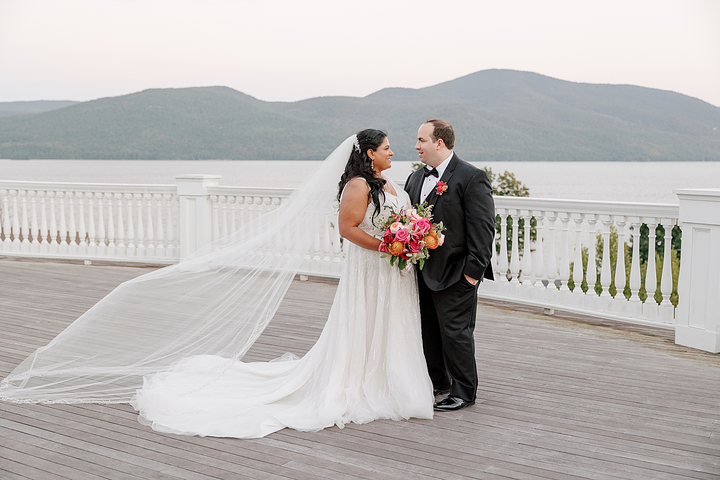 Bride and groom looking at each other on a deck with water and mountains in the background  | Image by Hope Helmuth Photography