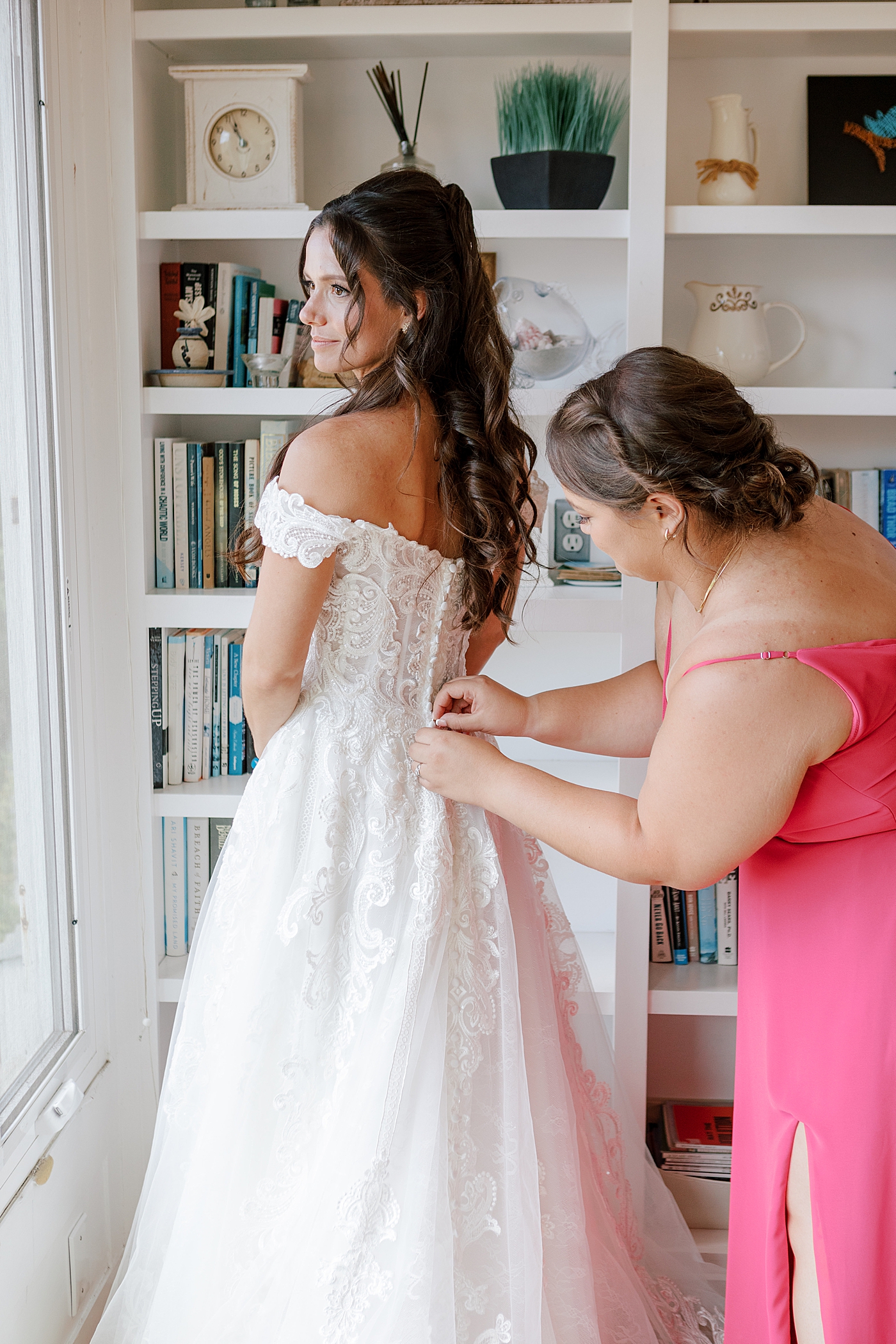 Bridesmaid in a pink dress buttoning the bride in her wedding dress in front of a bookshelf | Image by Hope Helmuth Photography