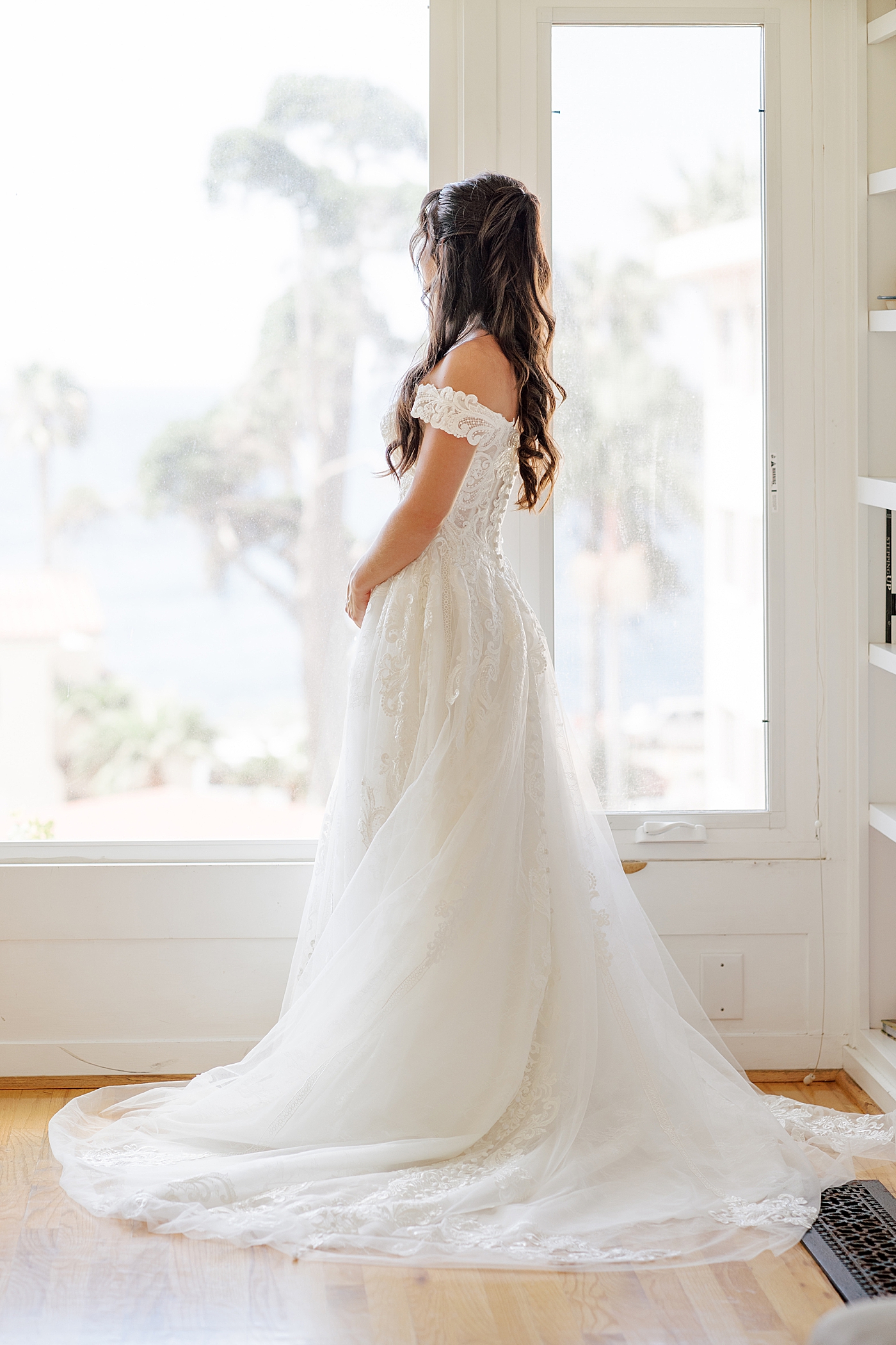 Full length image of a bride looking away from the camera and out a large, glass picture window | Image by Hope Helmuth Photography