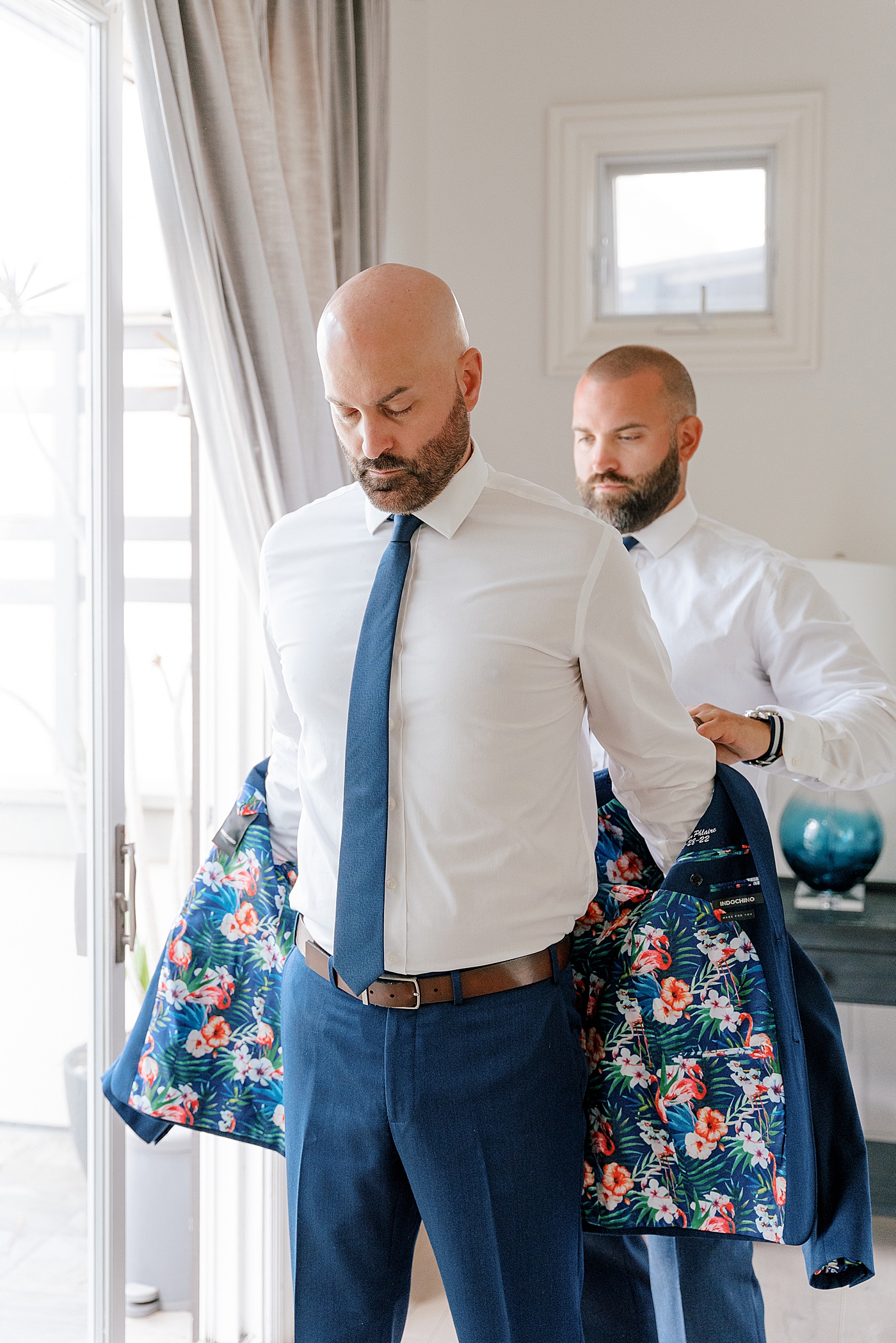 Groomsmen getting ready by helping each other put on navy jackets with bright, floral lining | Image by Hope Helmuth Photography