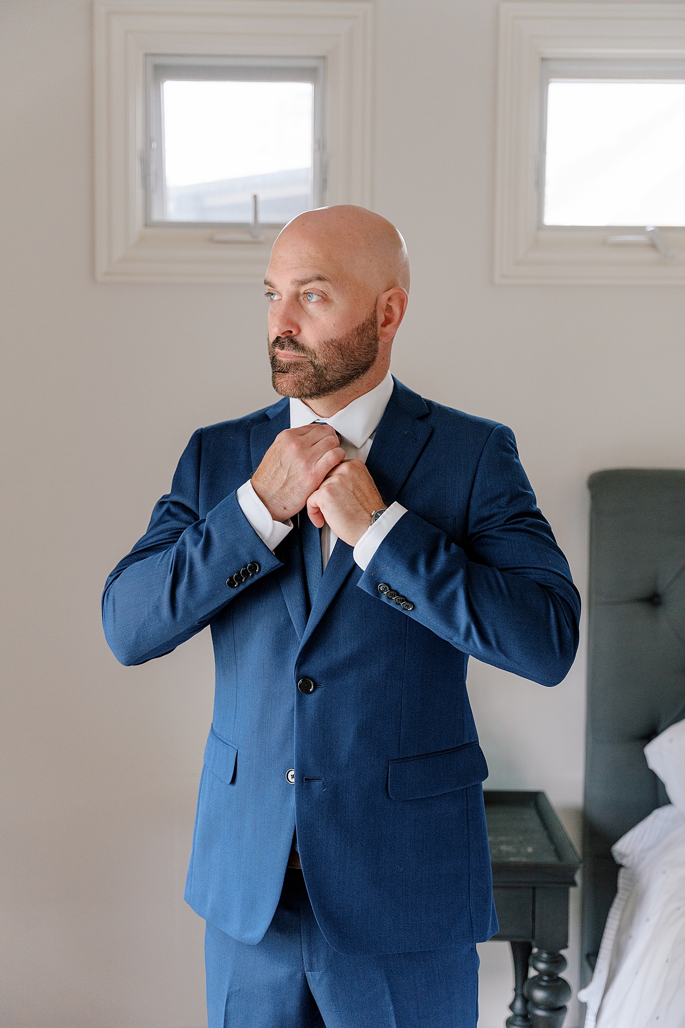 Groom in a navy suit looking towards window while fixing his tie | Image by Hope Helmuth Photography