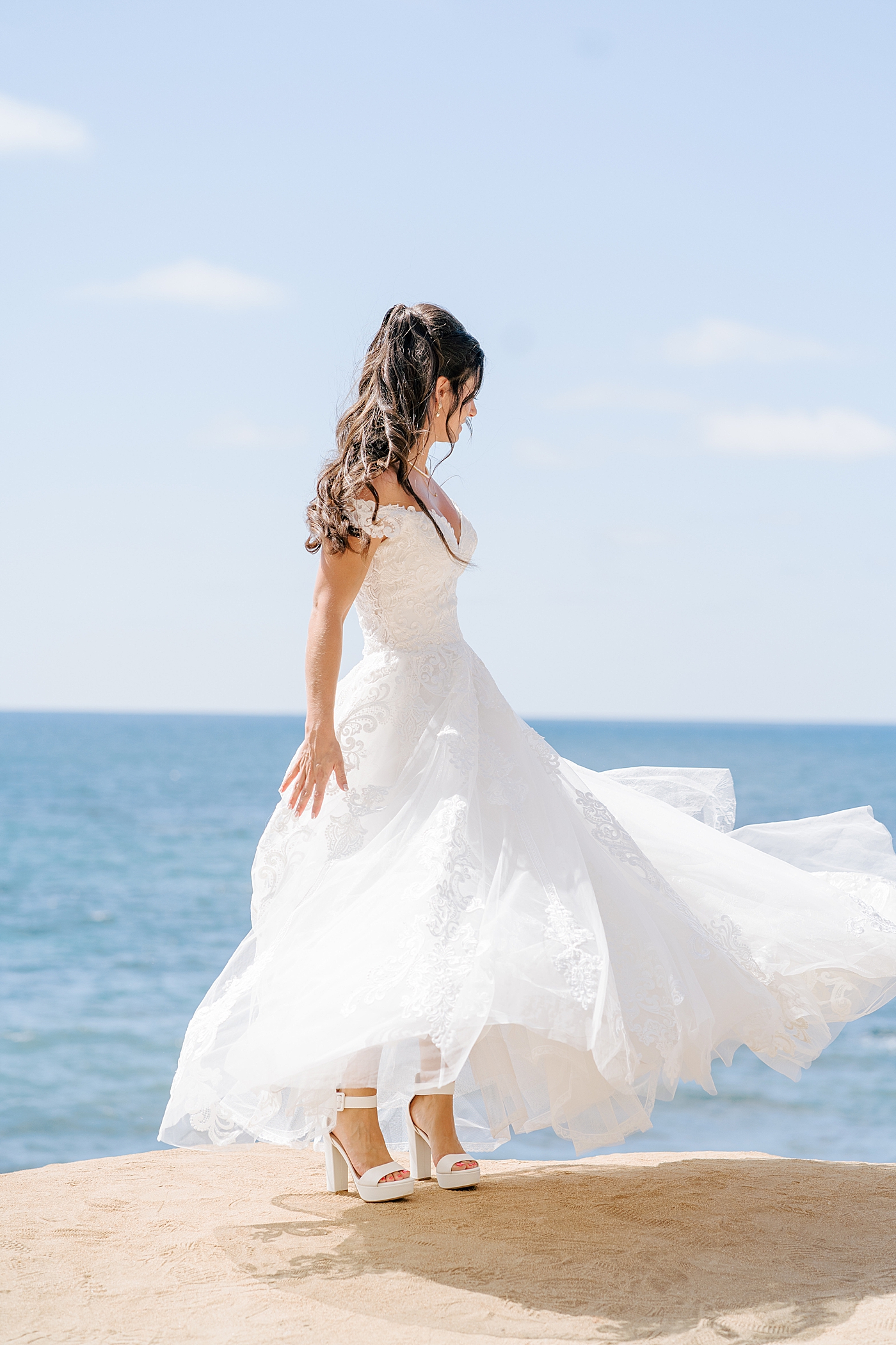 Bright photo of a bride oceanfront looking away from the camera while spinning in her dress | Image by Hope Helmuth Photography