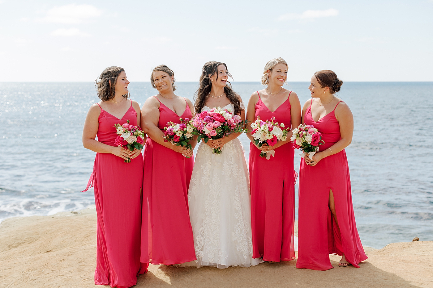Bride and bridesmaids in bright pink dresses on the beach by the water | Image by Hope Helmuth Photography