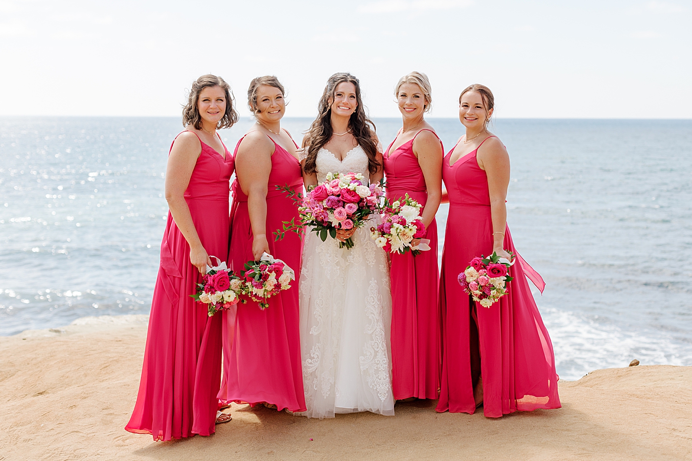 Bride and bridesmaids in hot pink looking at the camera smiling on the beach | Image by Hope Helmuth Photography
