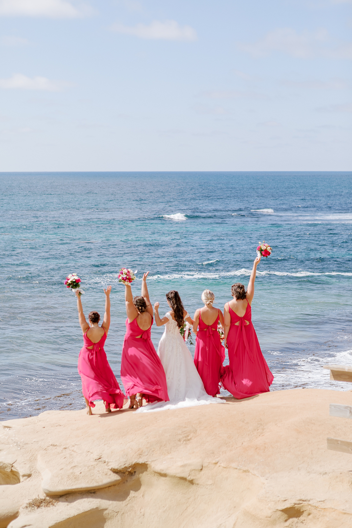 Bride and bridesmaids in hot pink holding bouquets up in celebration, facing away from the camera and photographed at a distance | Image by Hope Helmuth Photography