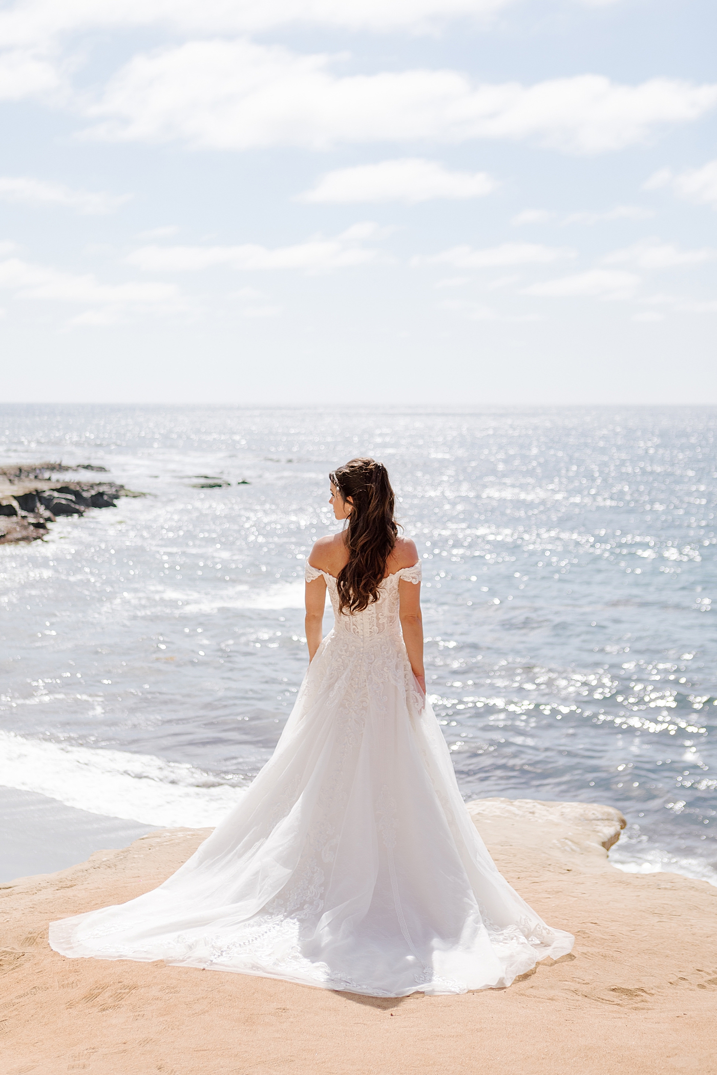 Bride standing oceanfront in backlit, full length photo displaying wedding dress | Image by Hope Helmuth Photography