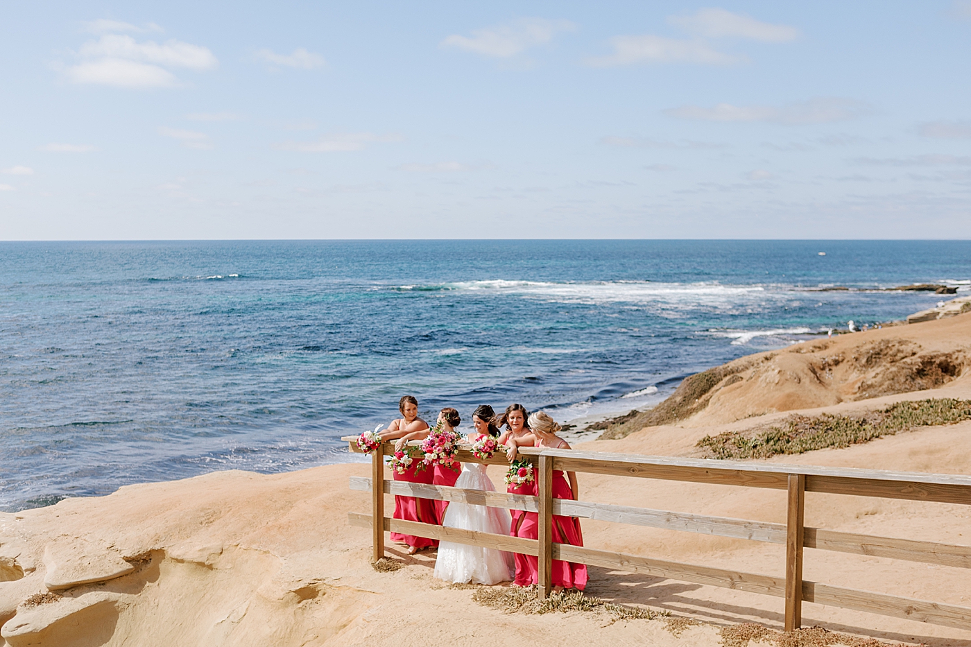Bride and bridesmaids in hot pink laughing and posing at a beach entrance with colorful bouquets | Image by Hope Helmuth Photography