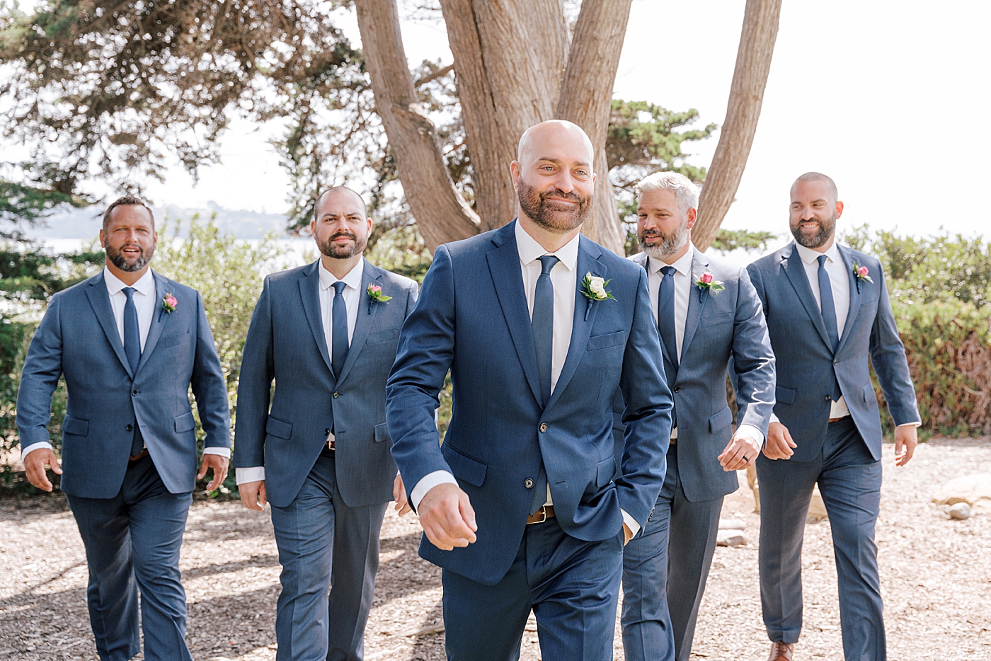 Groom and groomsmen in navy suits with one hand in a pocket walking towards the camera and smiling | Image by Hope Helmuth Photography