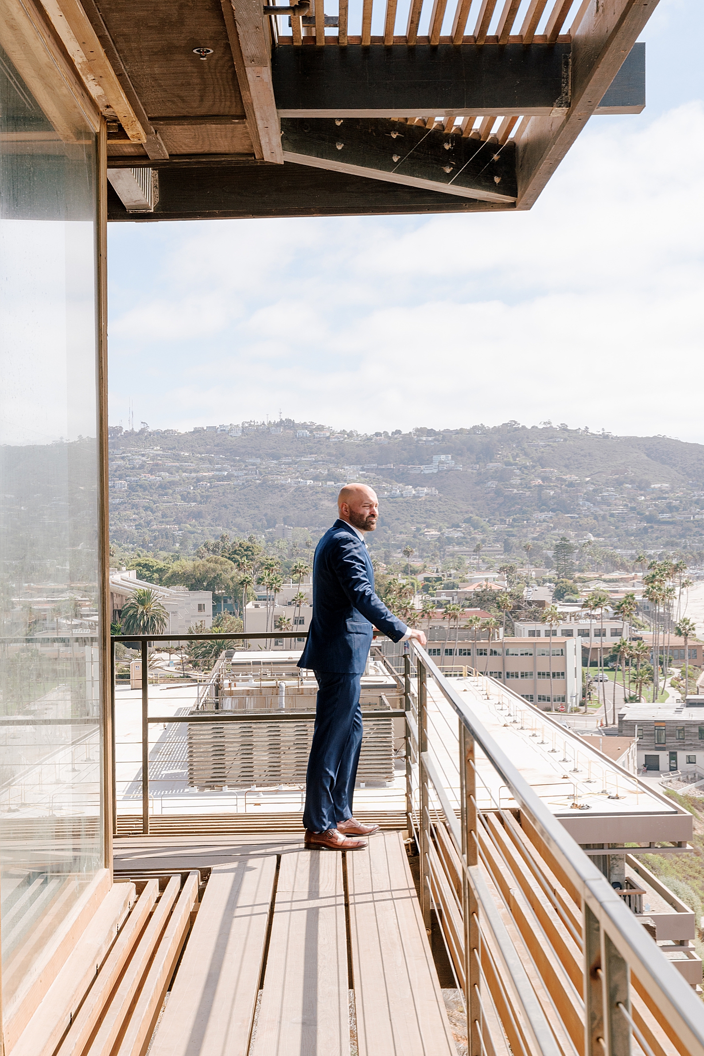 Groom in navy suit standing alone on the balcony looking out on a tropical, sunny city | Image by Hope Helmuth Photography