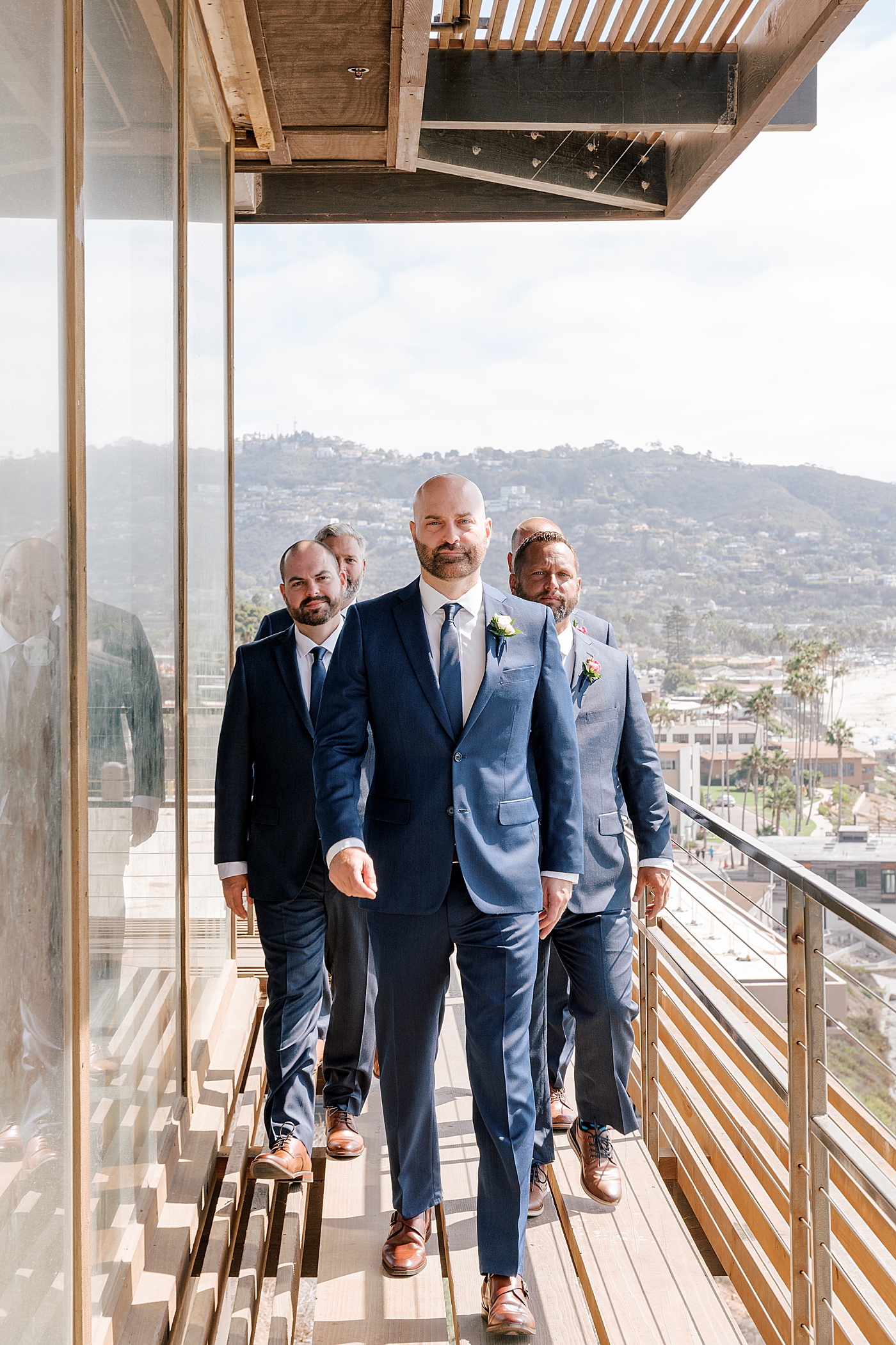 Groom and groomsmen in navy suits walking towards the camera on a balcony overlooking a sunny, tropical city | Image by Hope Helmuth Photography