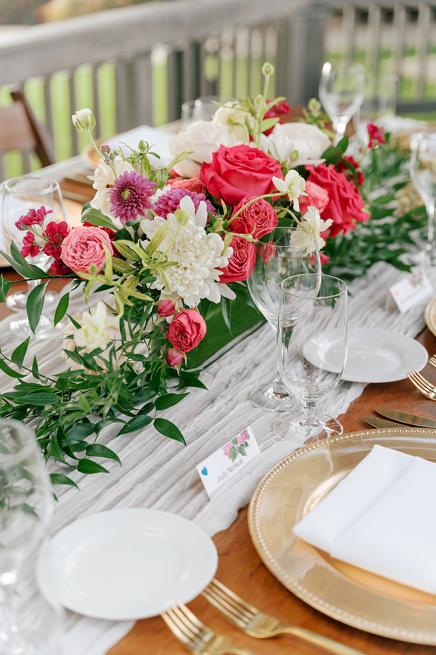 Detail of a wooden wedding dinner table with a white table runner, gold plates, and a colorful flower centerpiece | Image by Hope Helmuth Photography