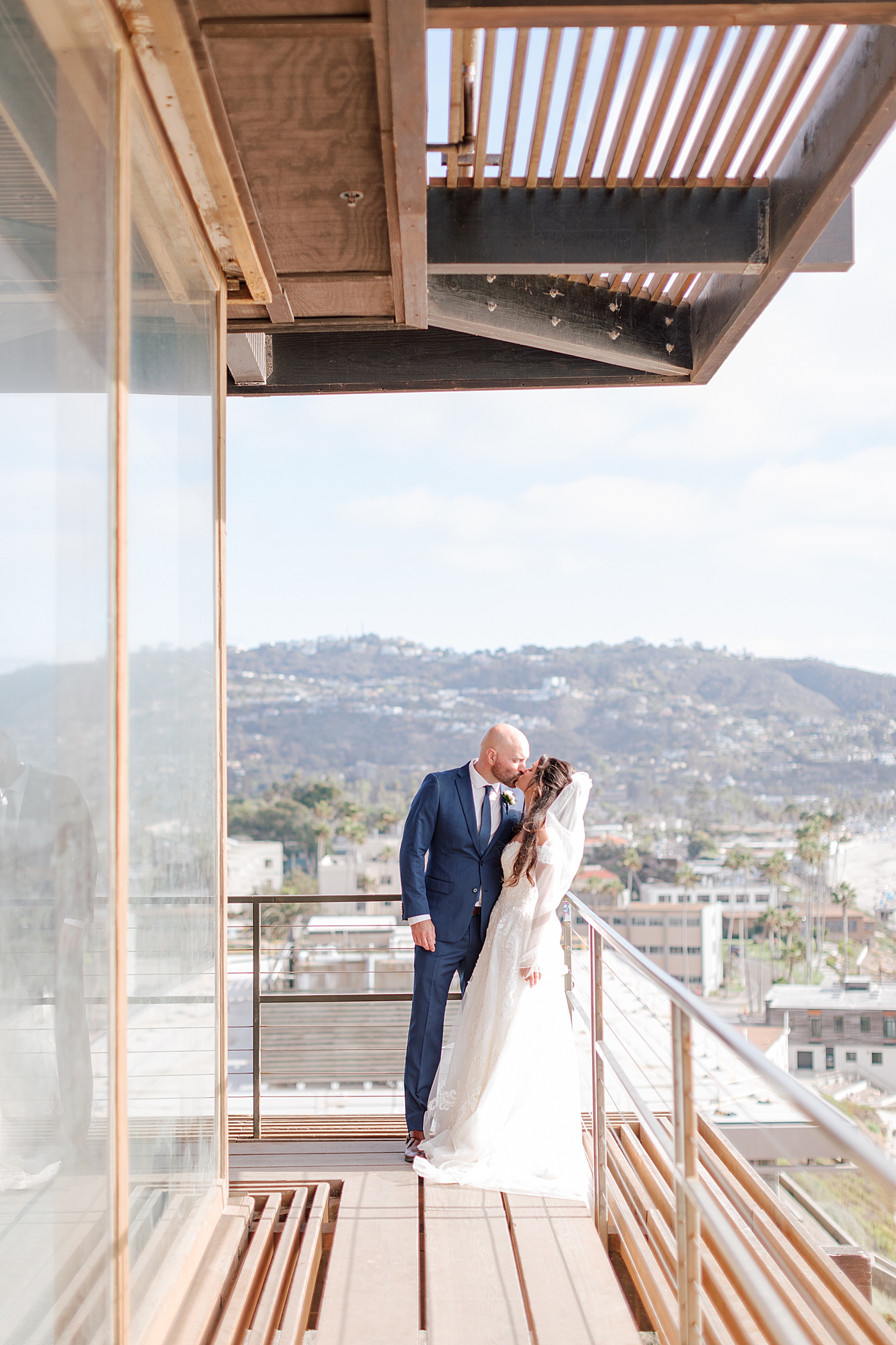 Groom in navy suit and bride in wedding dress standing closely and kissing on a wooden balcony that overlooks a tropical, sunny city | Image by San Diego Wedding Photographer Hope Helmuth