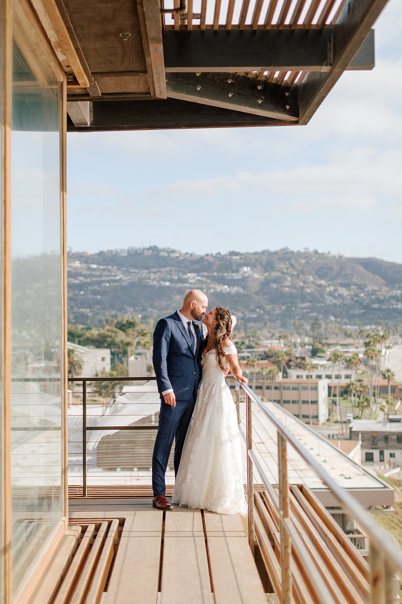 Groom in navy suit and bride in wedding dress standing closely and smiling at each other on a wooden balcony that overlooks a tropical, sunny city | Image by San Diego Wedding Photographer Hope Helmuth