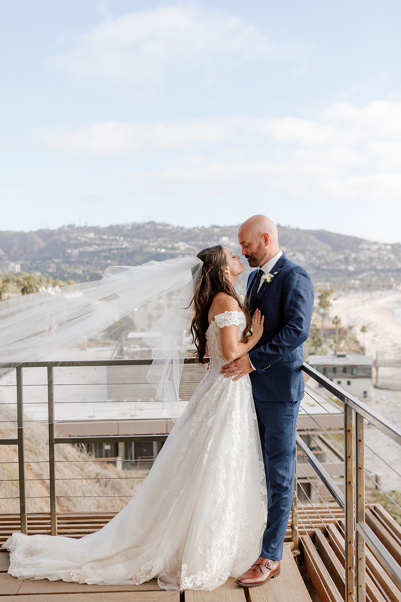 Close up of a groom in navy suit and bride in wedding dress standing closely and looking each other on a wooden balcony that overlooks a tropical, sunny city | Image by San Diego Wedding Photographer Hope Helmuth