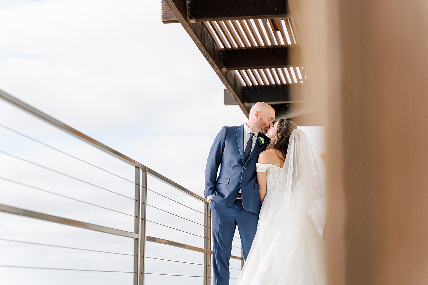 Groom in navy suit and bride in wedding dress standing closely and kissing on a wooden balcony | Image by Hope Helmuth Photography