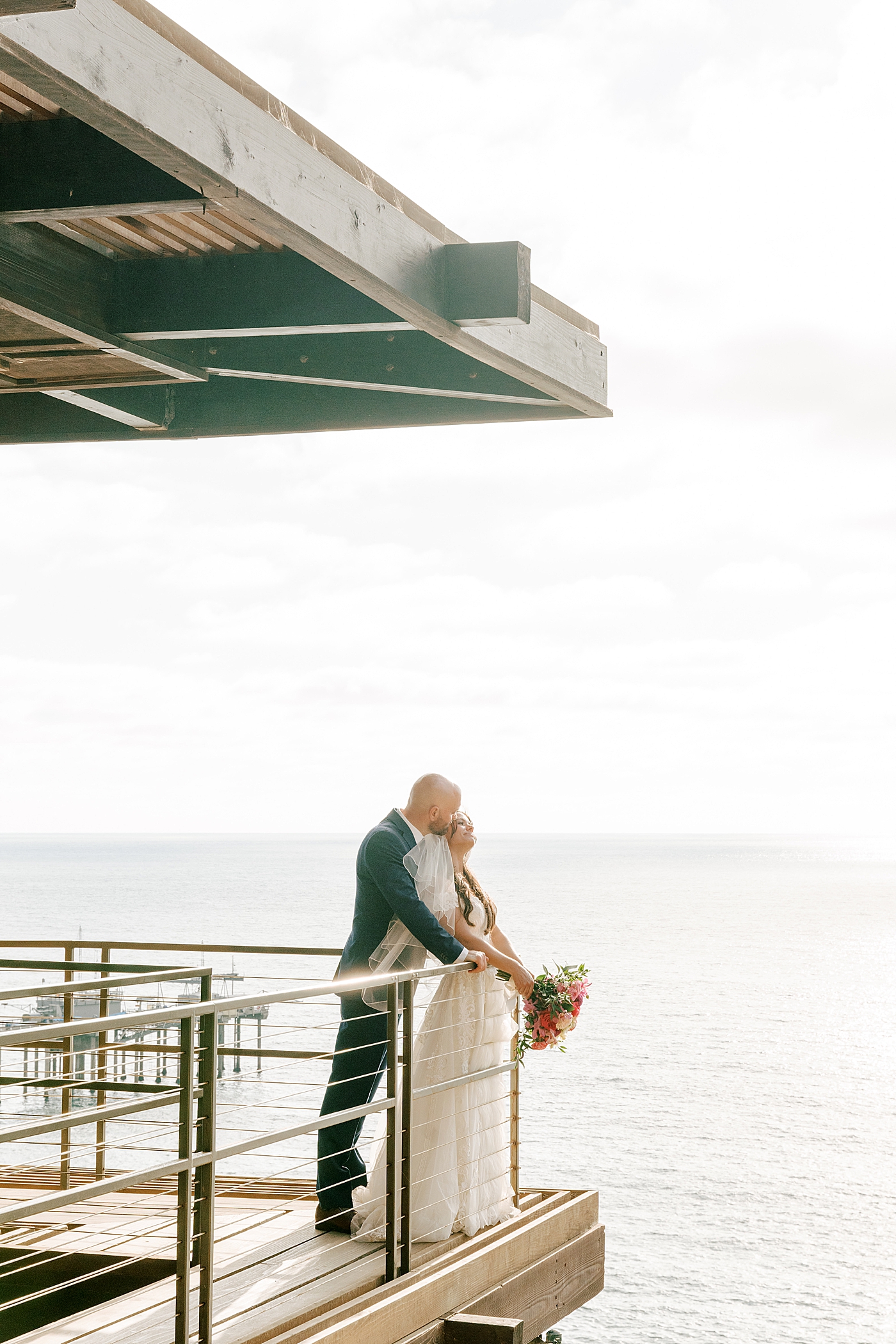 Groom in navy suit and bride in wedding dress standing closely on a wooden balcony that overlooks a tropical, sunny city | Image by Hope Helmuth Photography