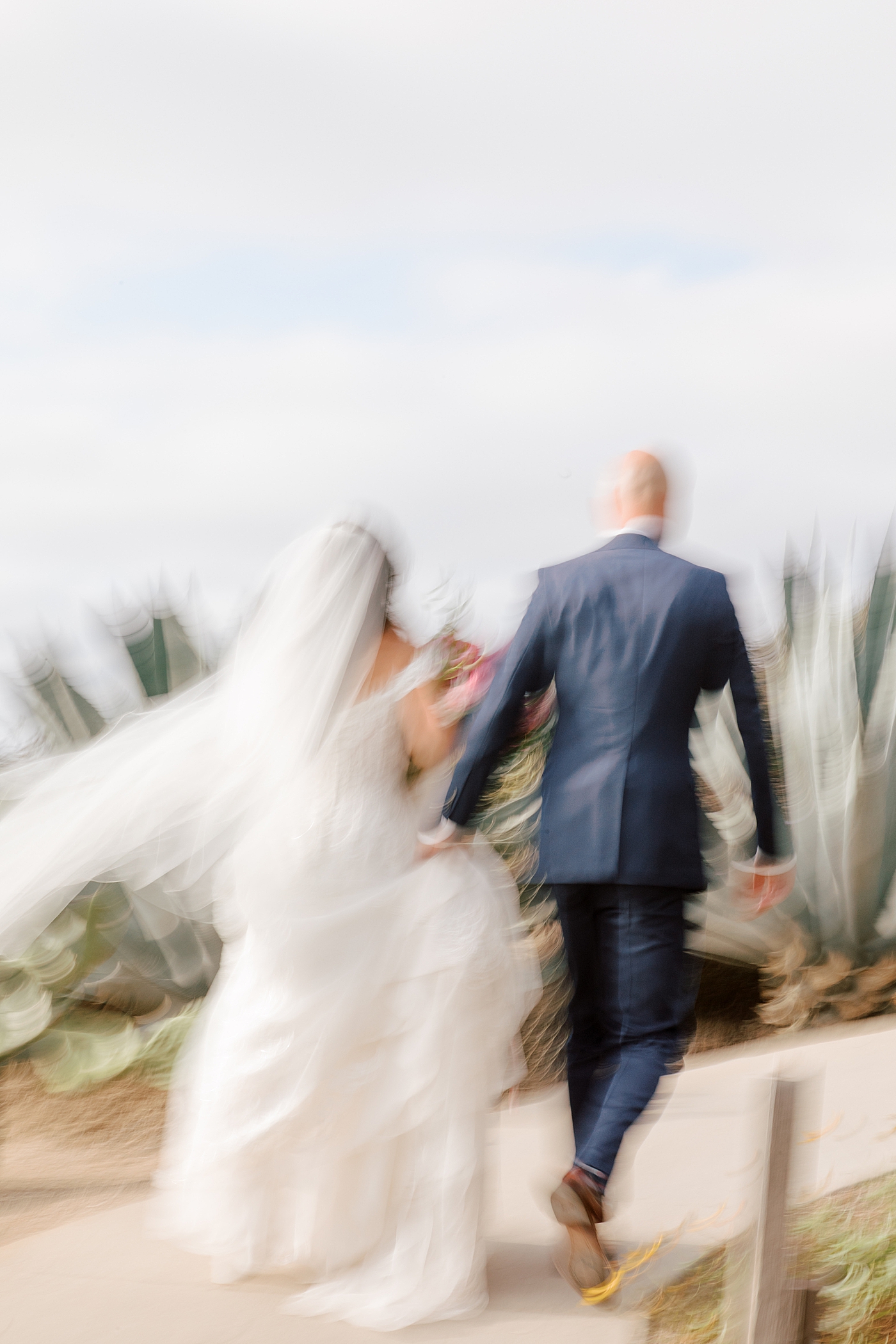 Blurry image of a groom in navy suit and bride in wedding dress holding hands while walking away from the camera on a seaside path | Image by Hope Helmuth Photography
