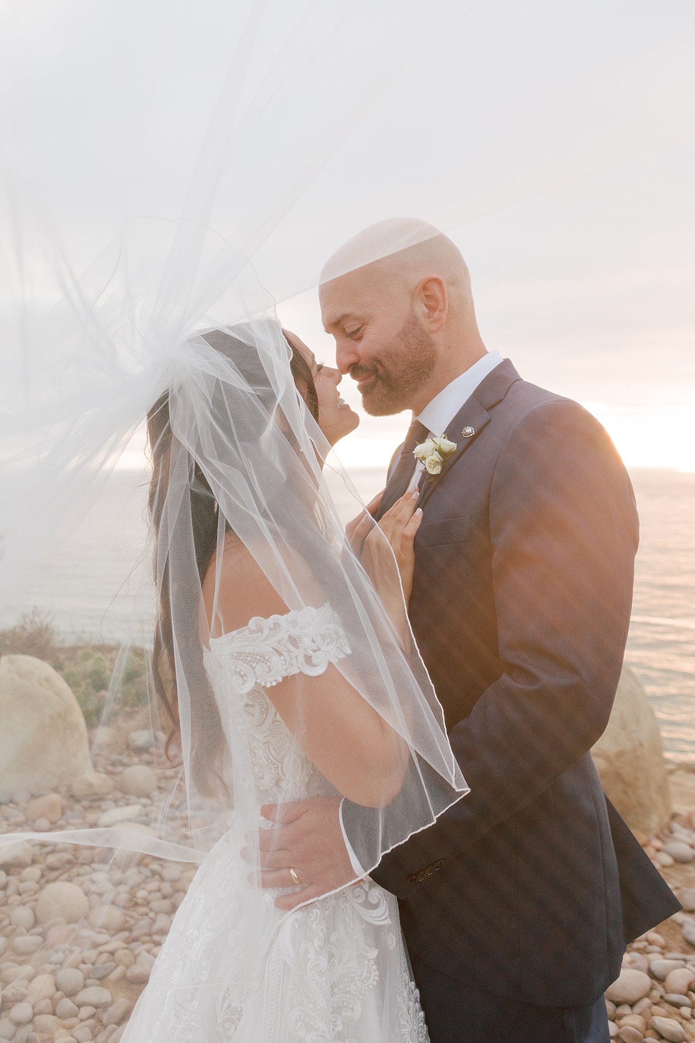Close up of a groom in navy suit and bride in wedding dress holding each other on a seaside path at sunset | Image by Hope Helmuth Photography