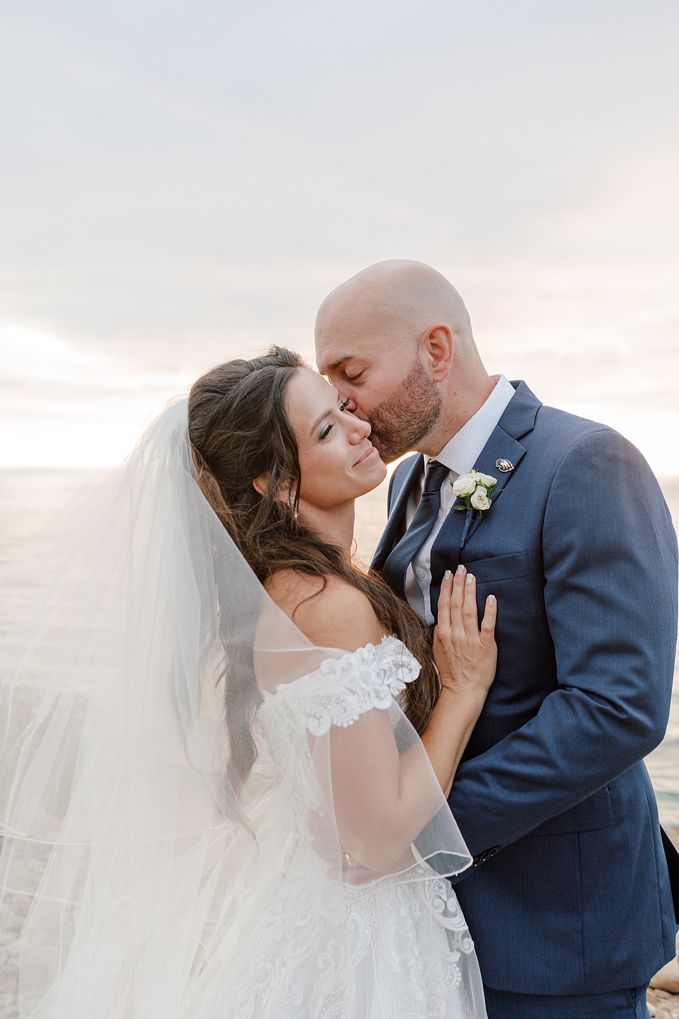 Close up of a groom in navy suit and bride in wedding dress holding each other with the groom kissing the bride on the cheek on a seaside path at sunset | Image by San Diego Wedding Photographer Hope Helmuth