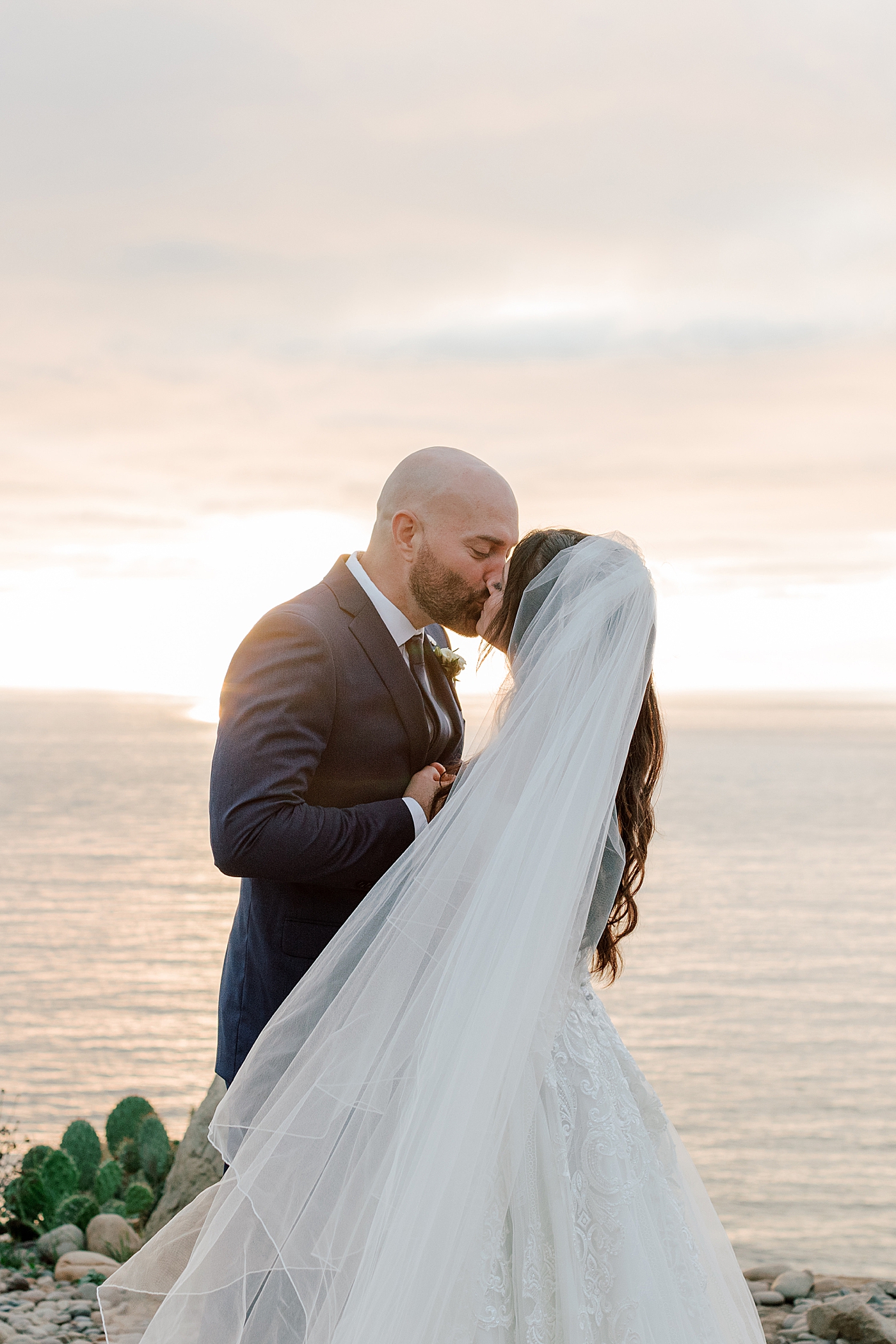 Groom in navy suit and bride in wedding dress holding each other and kissing on a seaside path at sunset | Image by San Diego Wedding Photographer Hope Helmuth