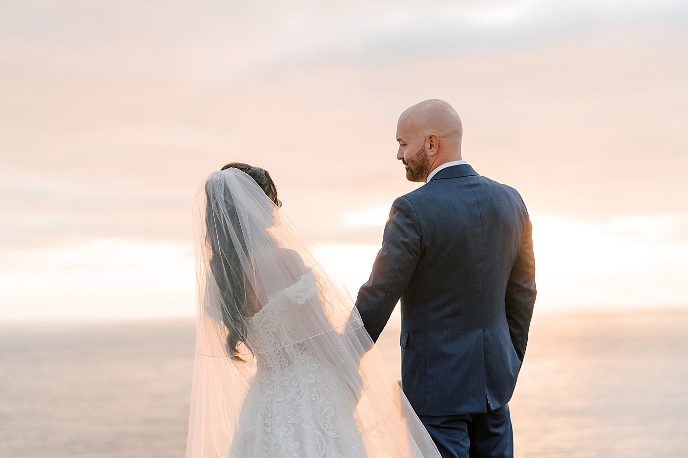 Groom in navy suit and bride in wedding dress facing away from the camera, looking at each other, and holding each other's hands on a seaside path at sunset | Image by Hope Helmuth Photography
