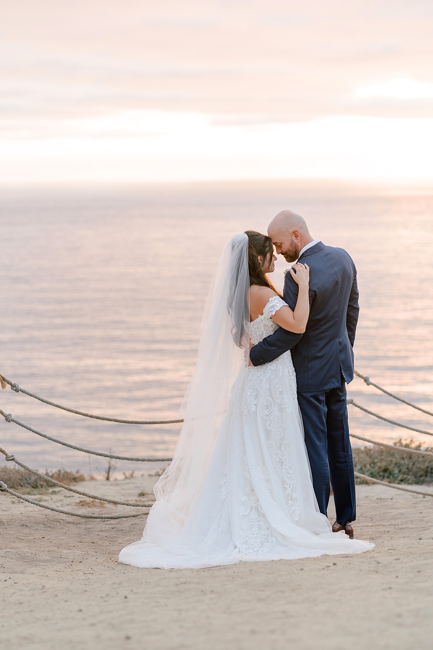 Groom in navy suit and bride in wedding dress holding each other closely on a seaside path at sunset | Image by San Diego Wedding Photographer Hope Helmuth