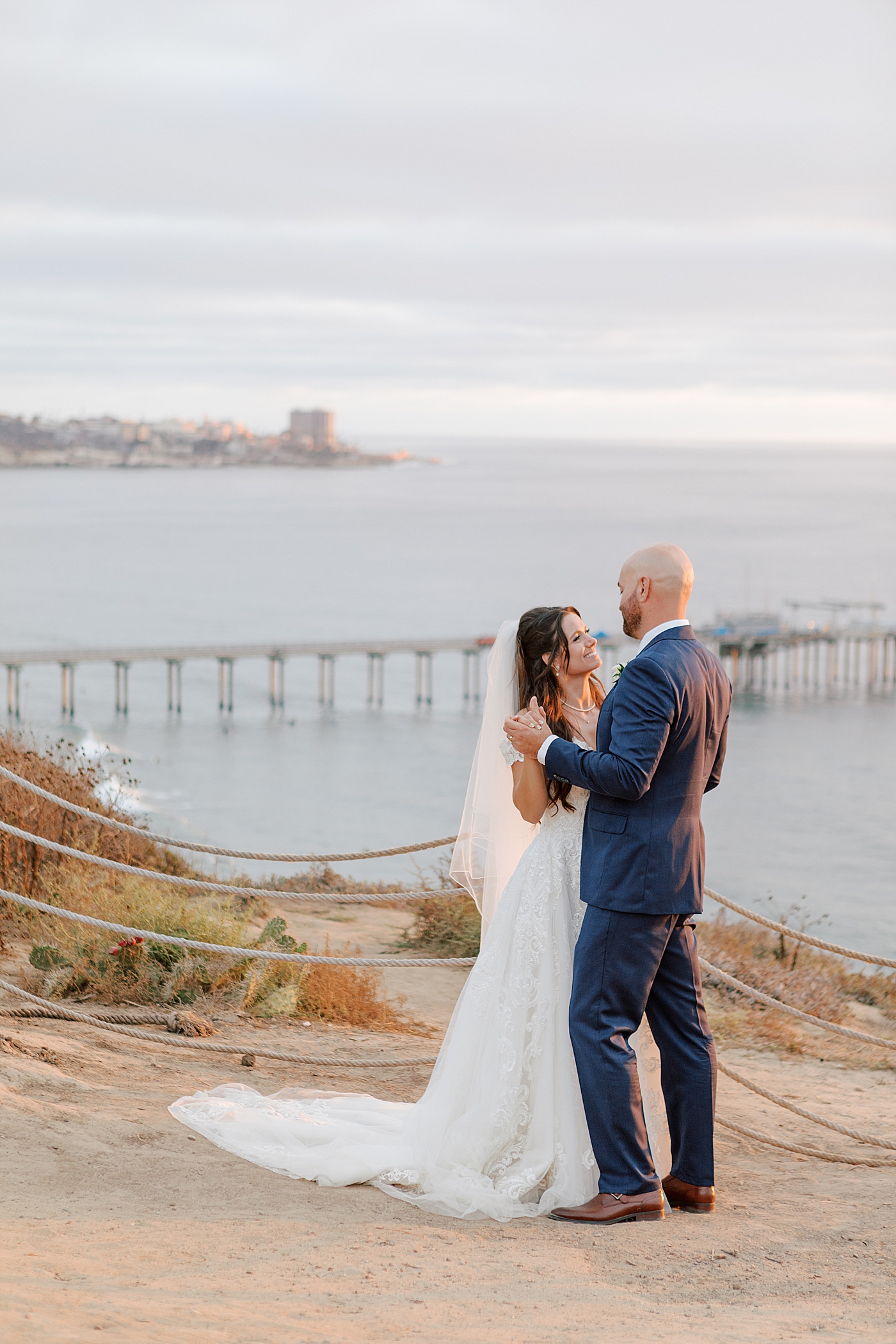 Groom in navy suit and bride in wedding dress holding each other and dancing on a seaside path at sunset | Image by Hope Helmuth Photography