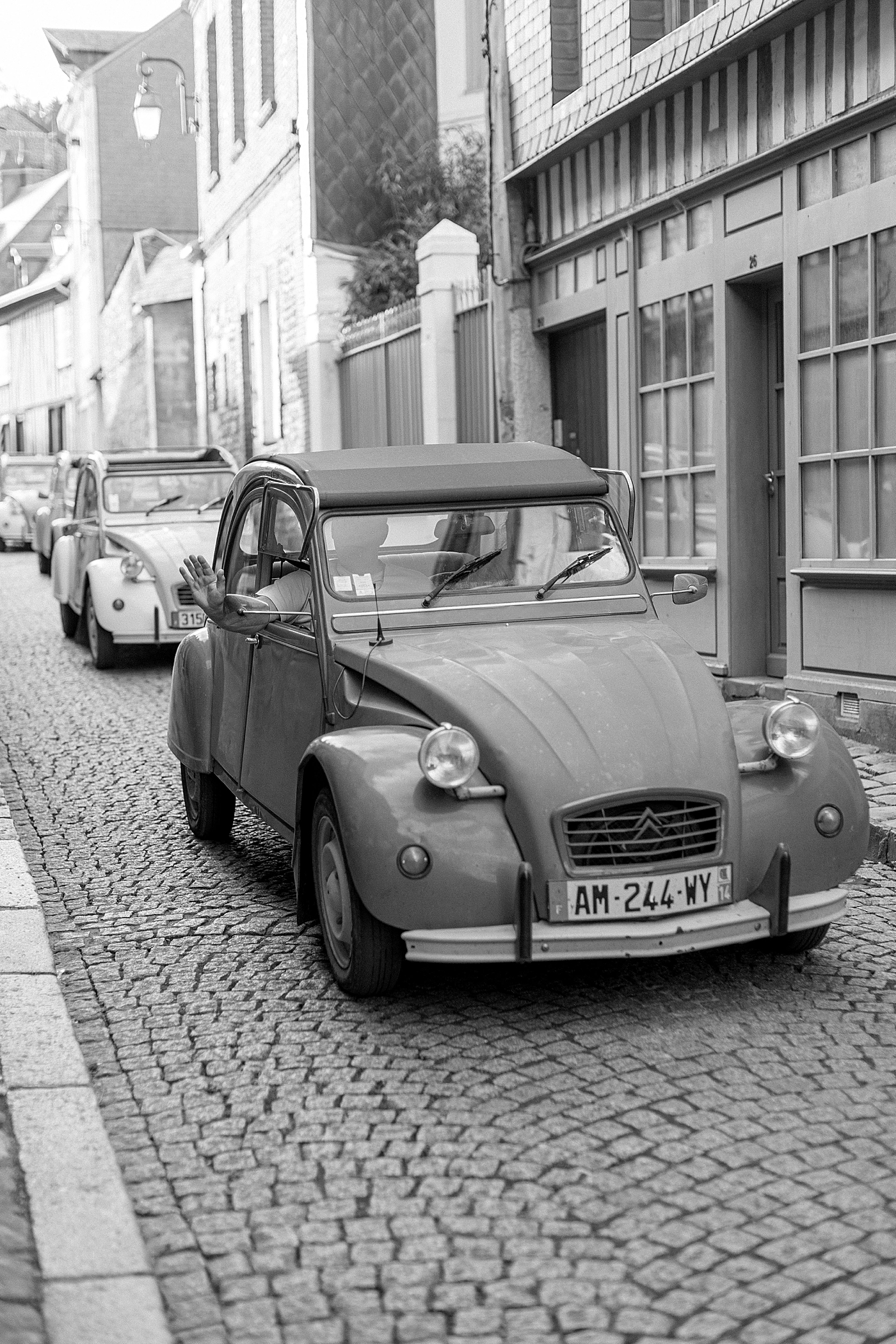 Black and white image of a driver waving out of his window while passing by on cobblestone streets during a trip with France Wedding Photographer Hope Helmuth Photography
